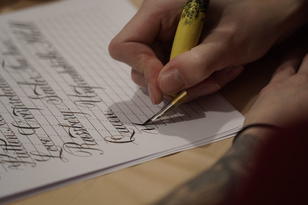 Close-up of a person's hand writing calligraphy with a yellow pen on lined paper.