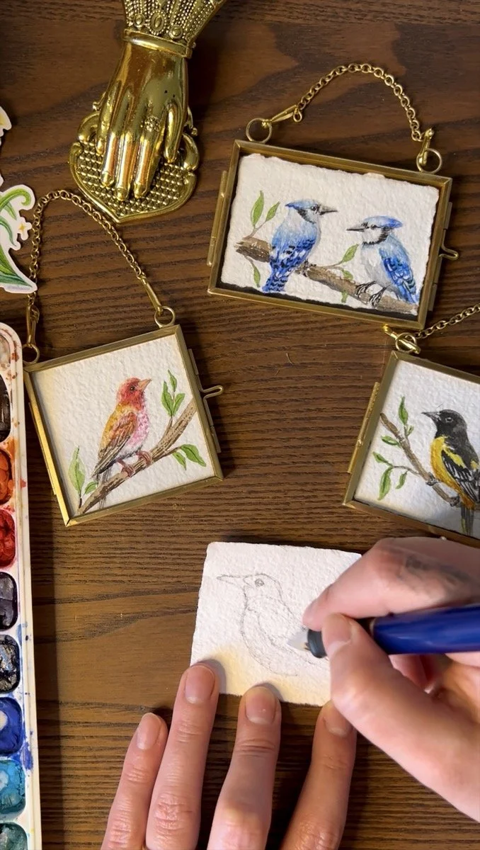 The image shows a person sketching a bird on a small piece of paper with a blue pen. The paper is held by one hand, and three painted bird illustrations in small golden frames with chains are on the wooden surface nearby. Additionally, there is a sma