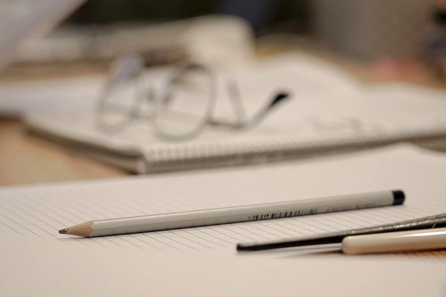 A close-up of a lined notebook with a gray mechanical pencil resting on it, and a black pen partially visible at the bottom edge. Blurred glasses and an open notebook are in the background.