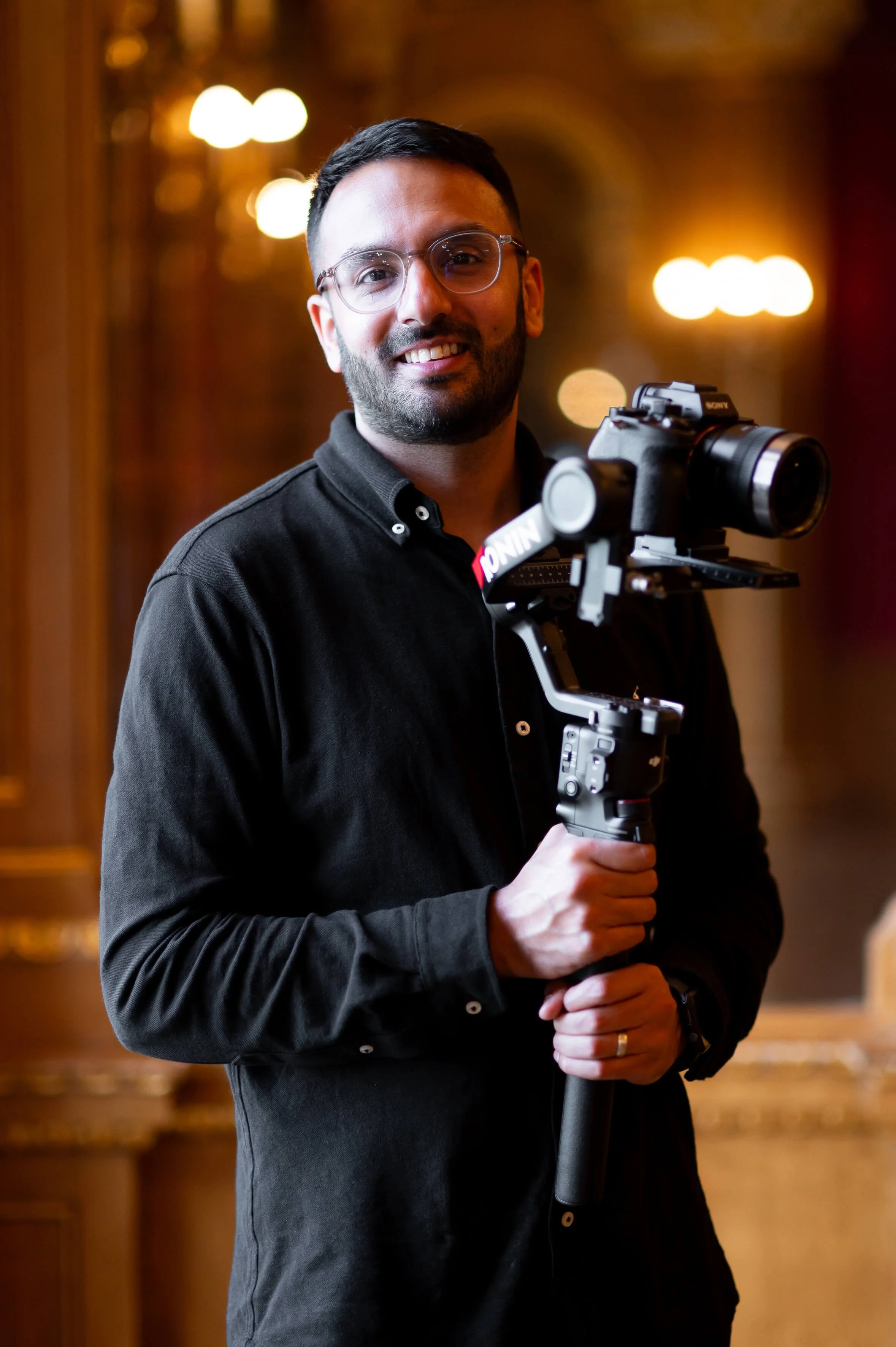 A man with glasses and a beard holding a professional camera on a stabilizer, smiling in a warmly lit indoor setting with blurred lights in the background.