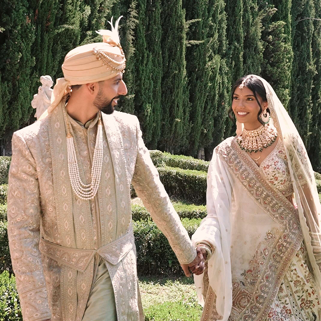 A bride and groom in traditional Indian wedding attire holding hands outdoors, surrounded by greenery and tall trees.