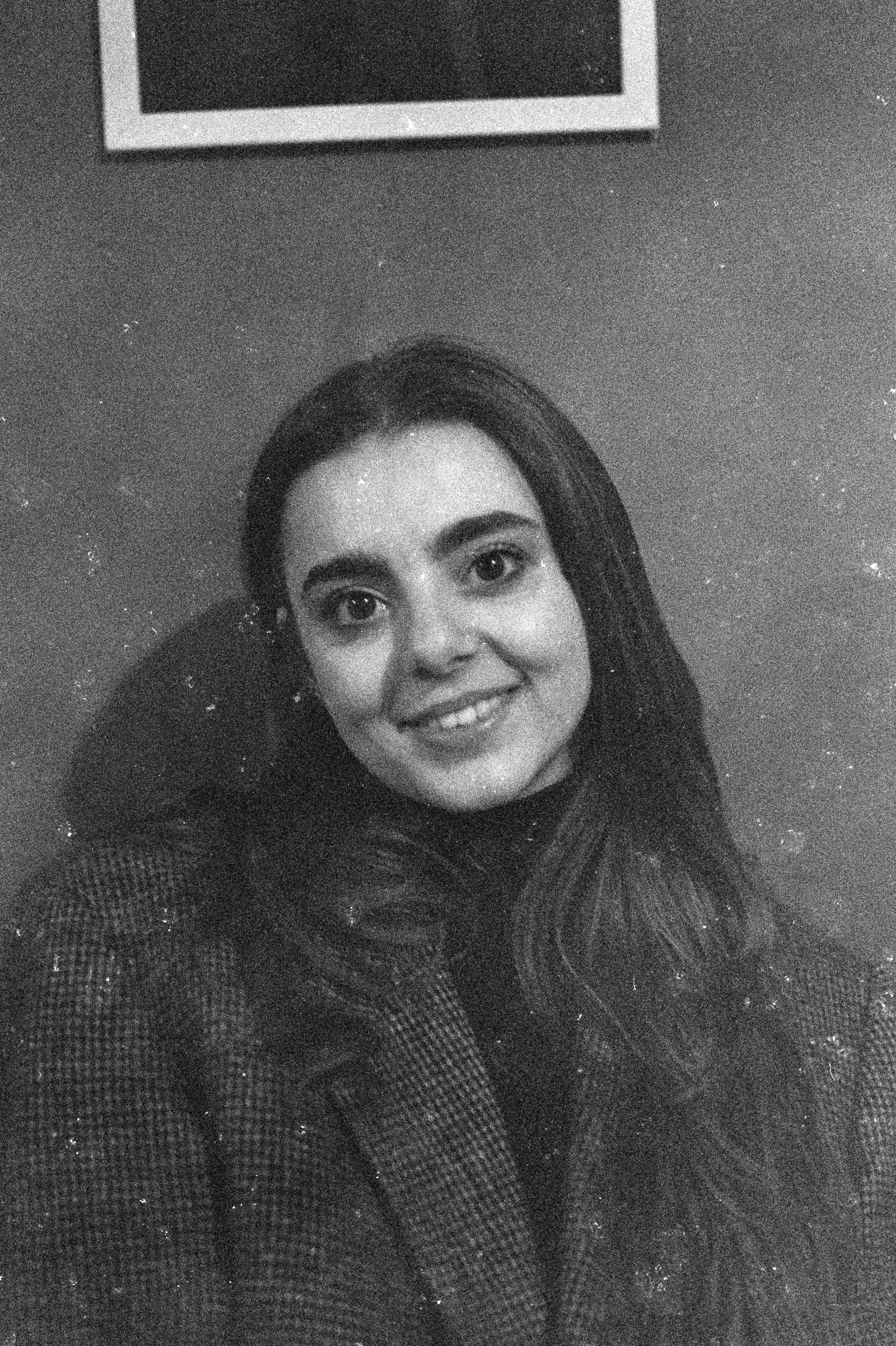 Black-and-white photo of a young woman with long hair smiling at the camera, standing against a plain wall with a framed picture above her head.