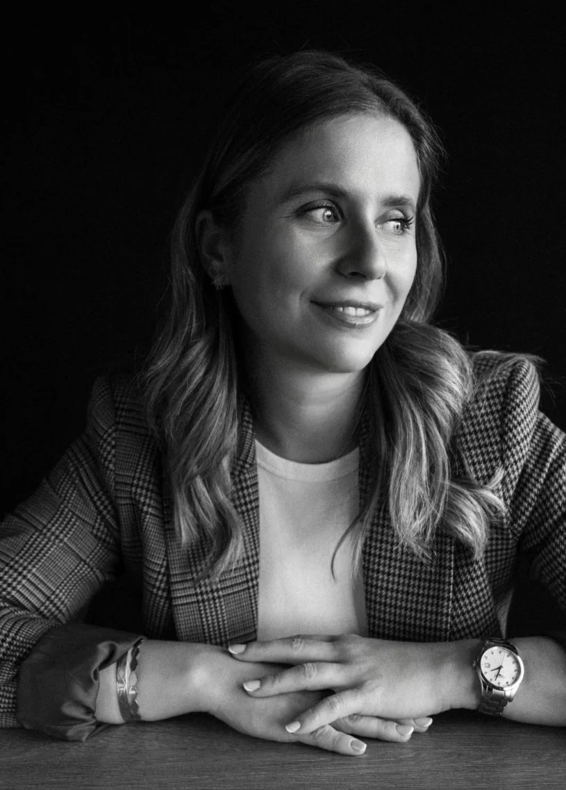 Black and white portrait of a woman with wavy hair, smiling, wearing a checked blazer, a watch, and jewelry, sitting with folded hands on a table.