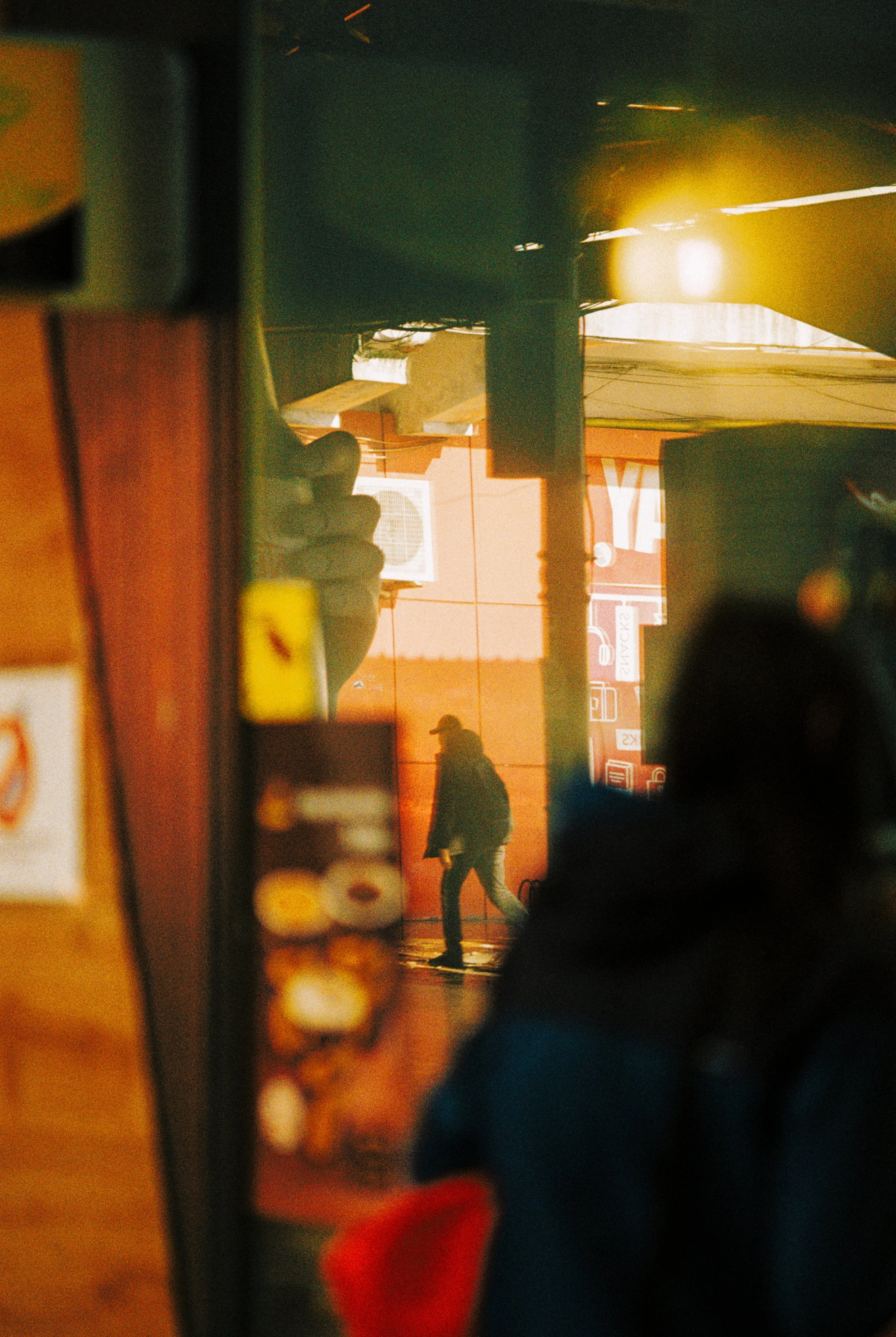 A person walking outside at night, seen through a reflective surface of a window or mirror, with street lights and signs in the background.