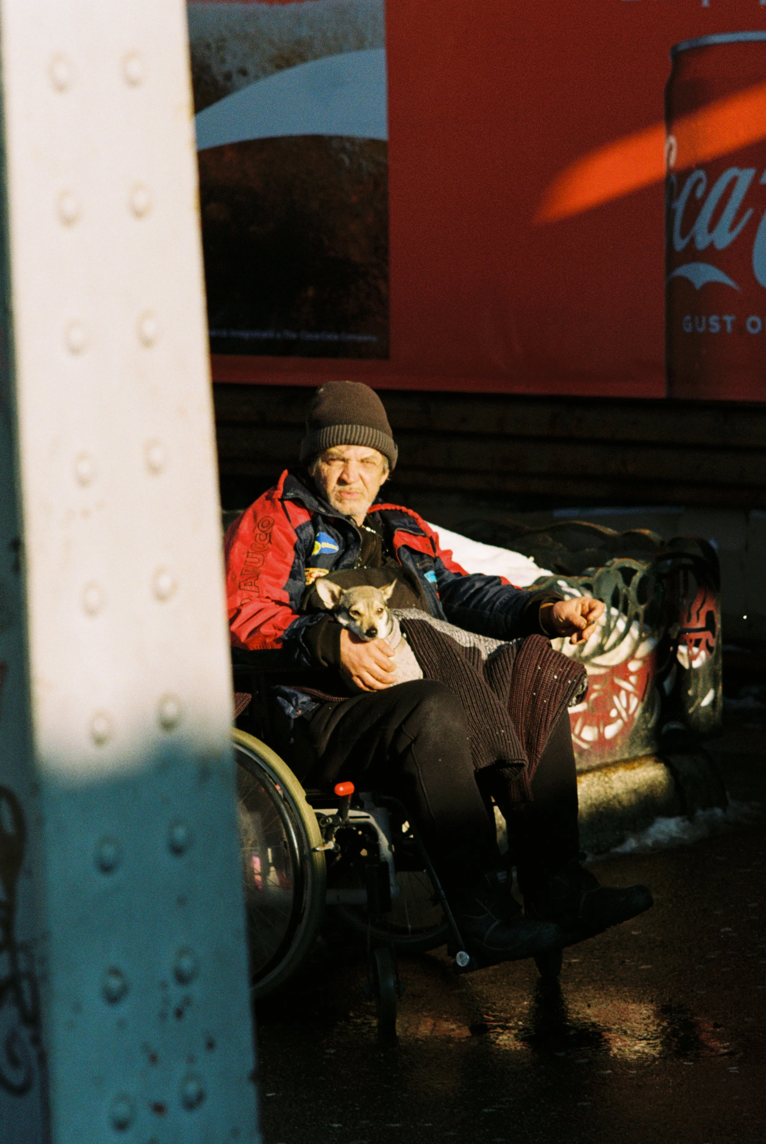 An elderly man in a wheelchair sitting on a bench outdoors, holding a small dog, with a large Coca-Cola advertisement in the background.