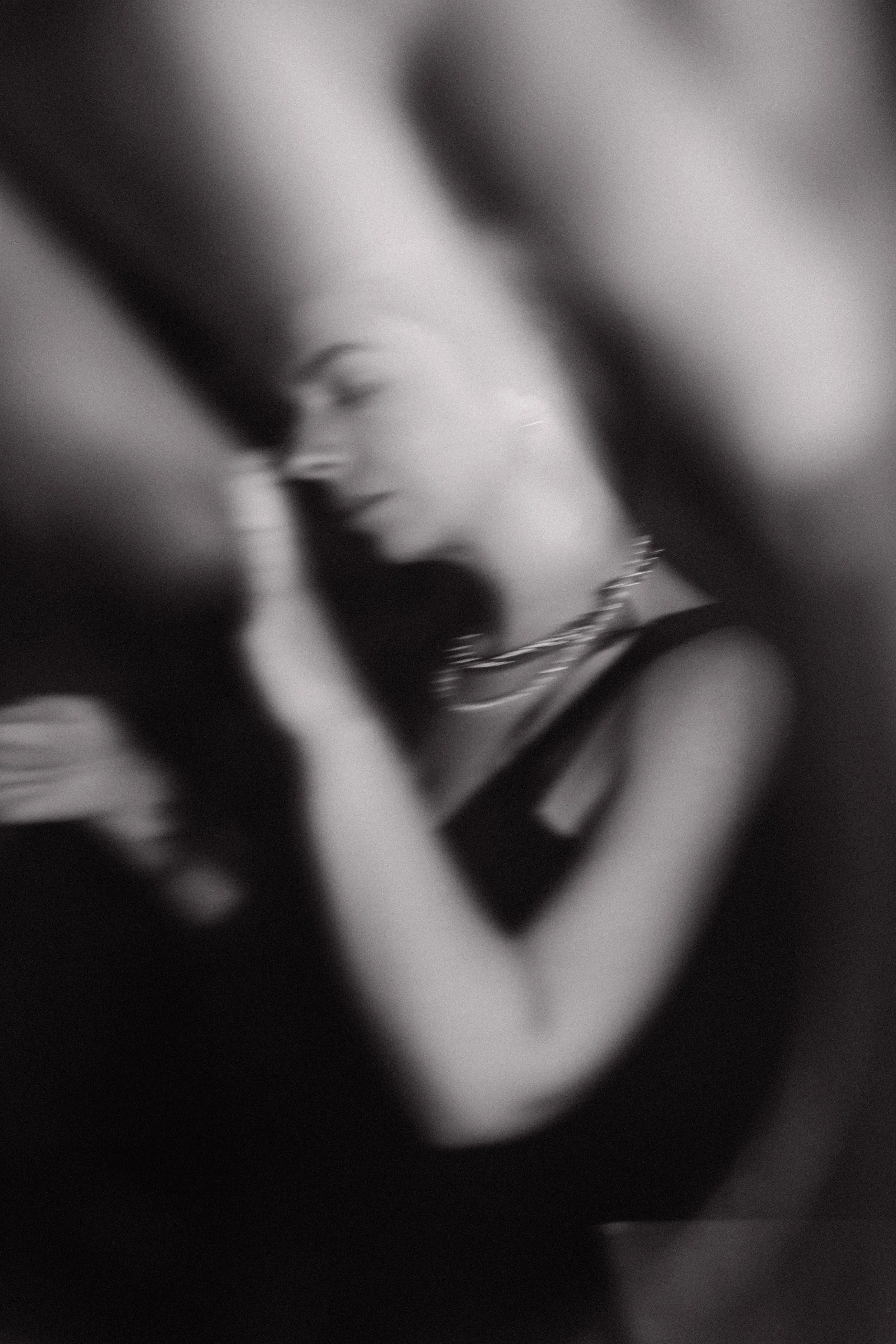 A blurry black-and-white photo of a woman with necklaces, possibly holding her face or head with her hand in a close-up shot.
