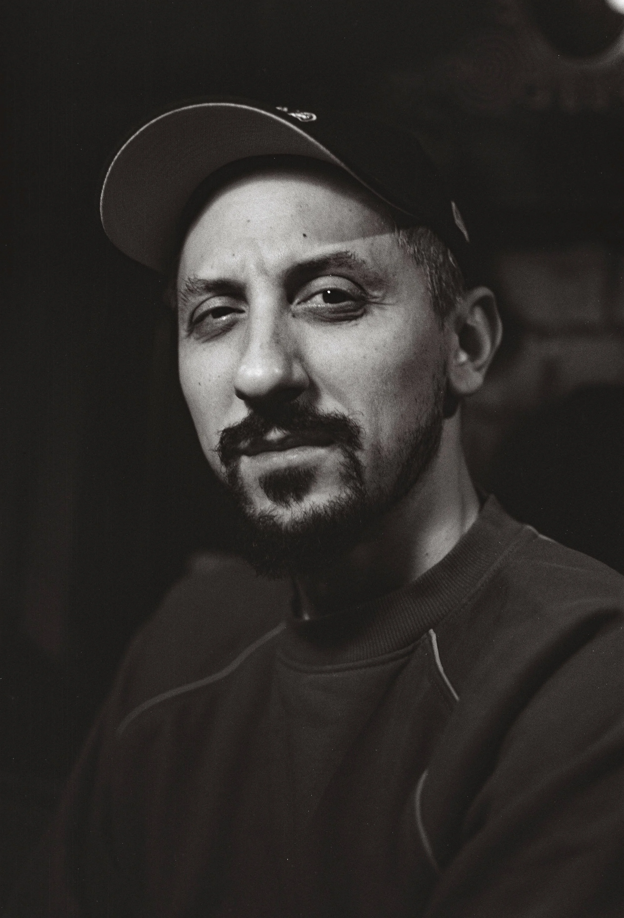 Black and white portrait of a man wearing a baseball cap, with a beard and mustache, looking at the camera.