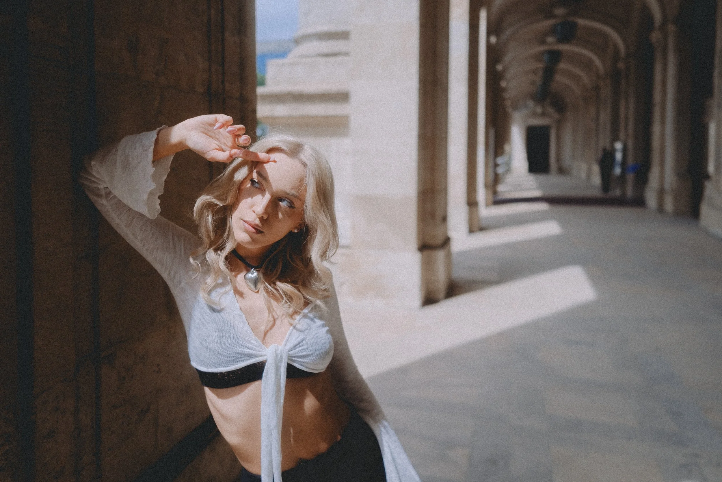 Young woman with blonde hair leaning against a stone pillar in an arched corridor, looking into the distance and shielding her eyes from the sun with her hand.