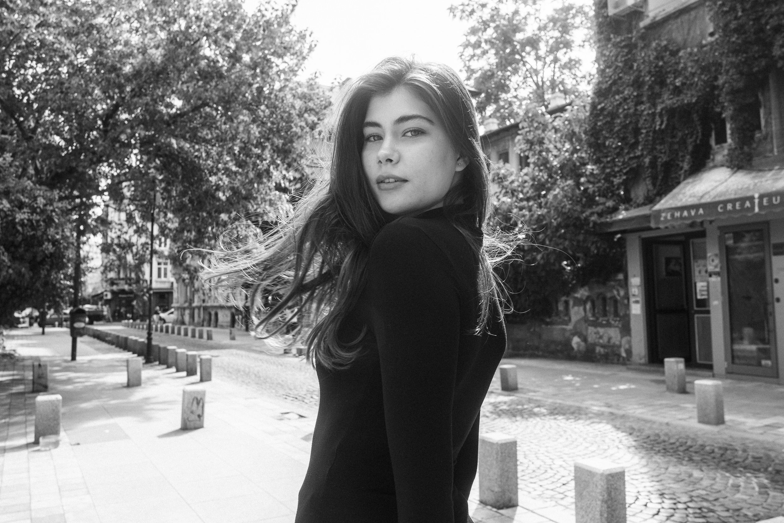 Black and white photo of a young woman with long hair, wearing a dark top, standing on a cobblestone street with trees and buildings in the background.
