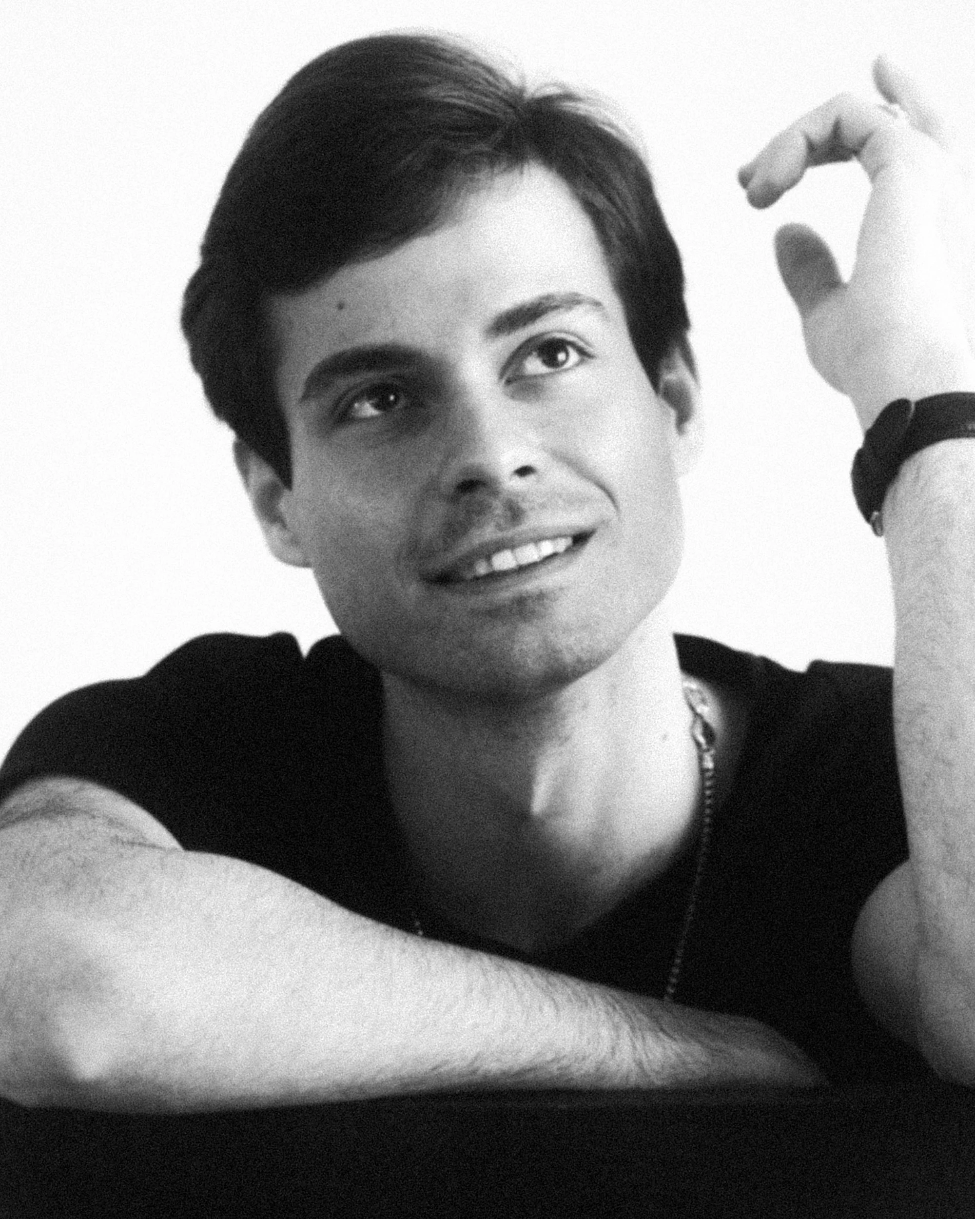 Black and white portrait of a young man with dark hair, smiling and looking slightly upward, wearing a black t-shirt and a watch on his left wrist.