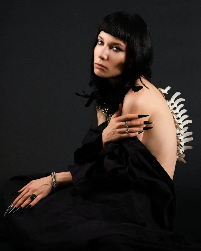 A woman with black hair and bold makeup, wearing black clothing and gothic jewelry, sitting with one shoulder exposed and a decorative spine accessory behind her, set against a dark background.