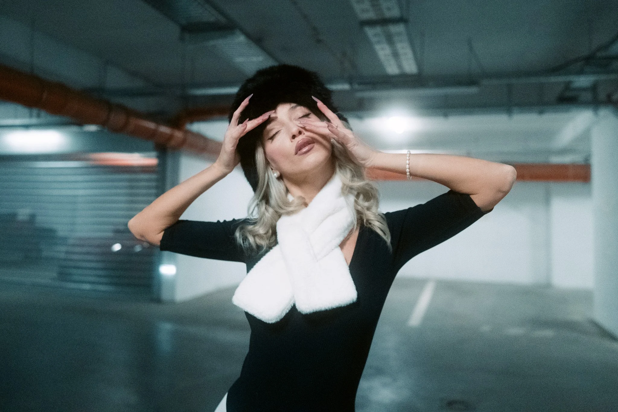A woman with blonde hair and a black hat, holding her head with both hands, appears distressed or overwhelmed, in an indoor parking garage.