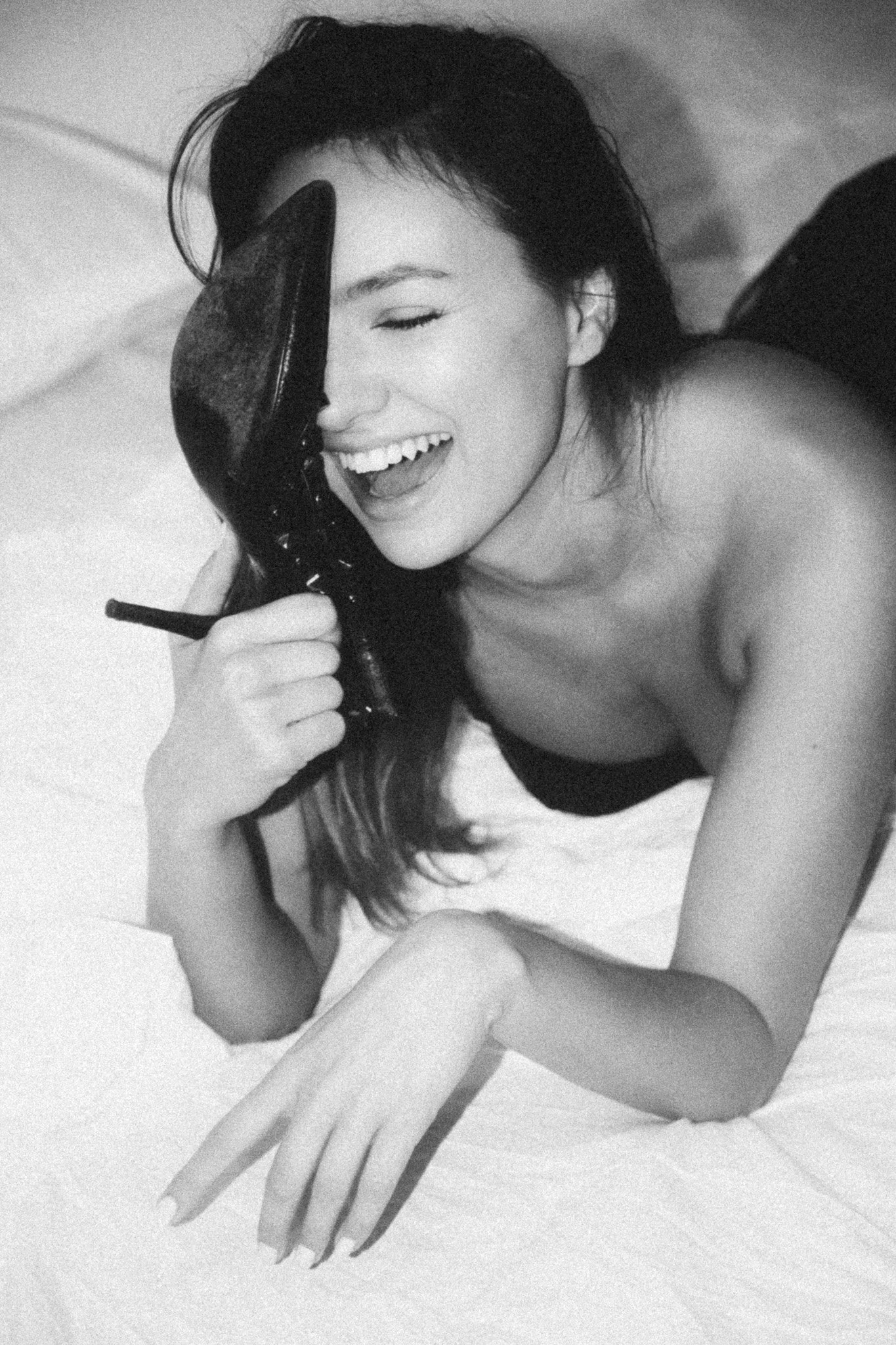 A woman with long dark hair laughing, holding a shoe to her face, lying on a bed, in black and white.