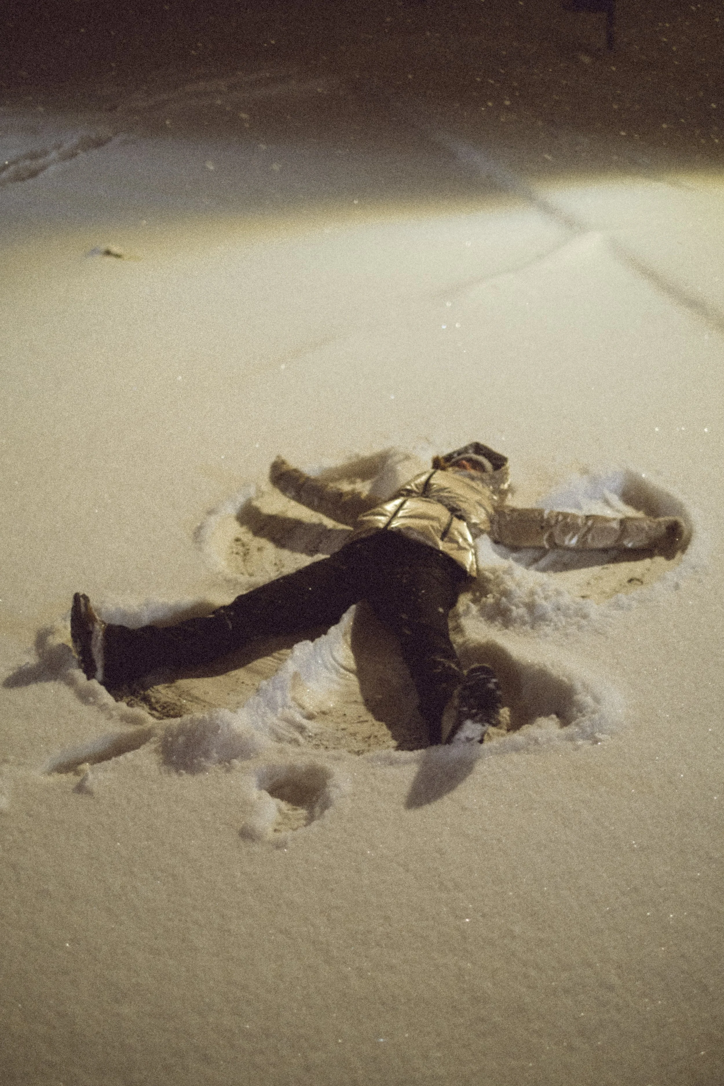 Child lying on snow, making snow angel, nighttime, wearing a shiny jacket and dark pants.