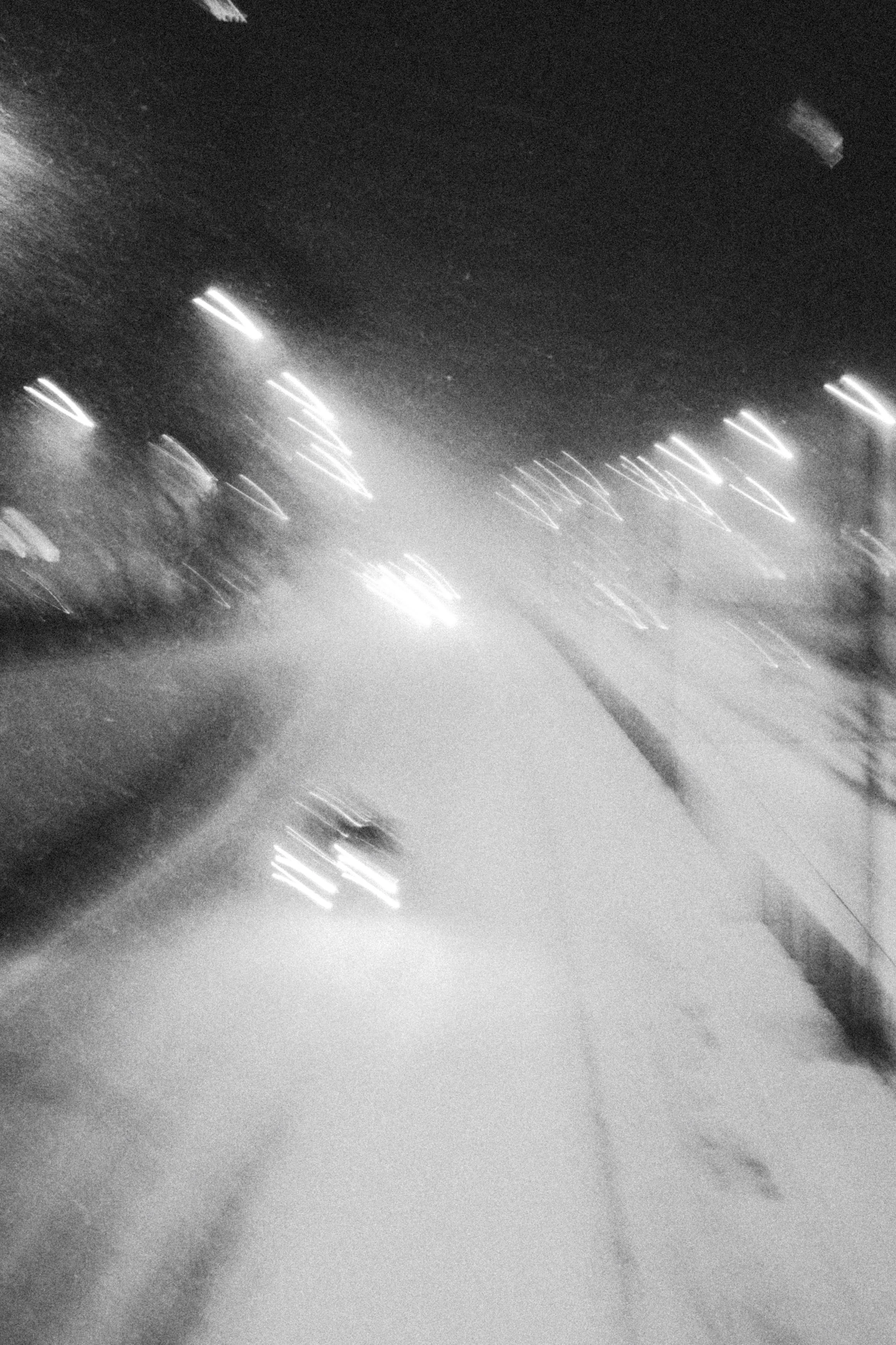 A black and white photo of a wet street at night, with blurry headlights and streetlights, and some snow on the ground.
