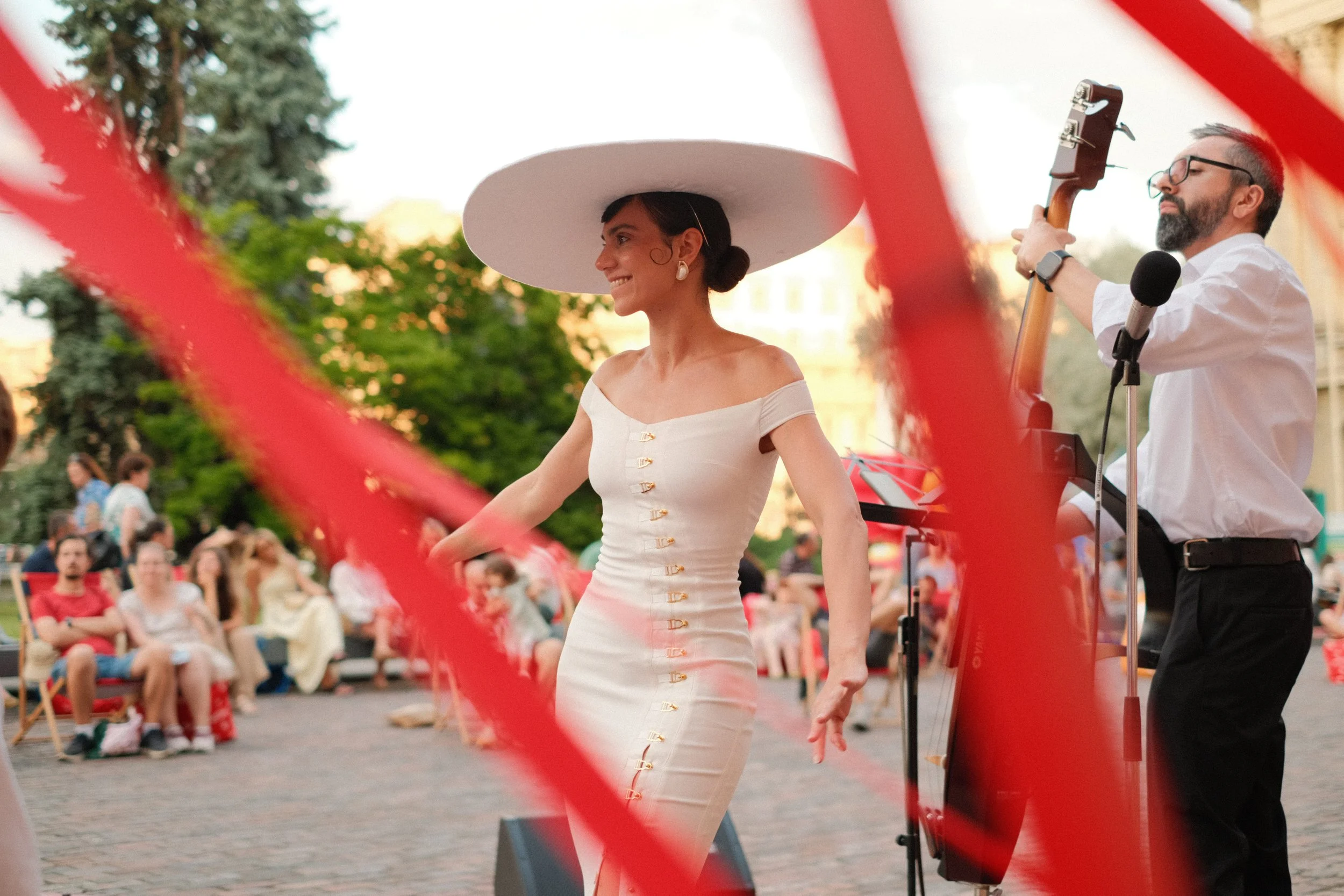 A woman dressed in a white, off-the-shoulder dress with gold buttons, wearing a large white wide-brimmed hat, is performing a dance in an outdoor plaza during a public event. A man playing a guitar is next to her, and a crowd is watching in the backg
