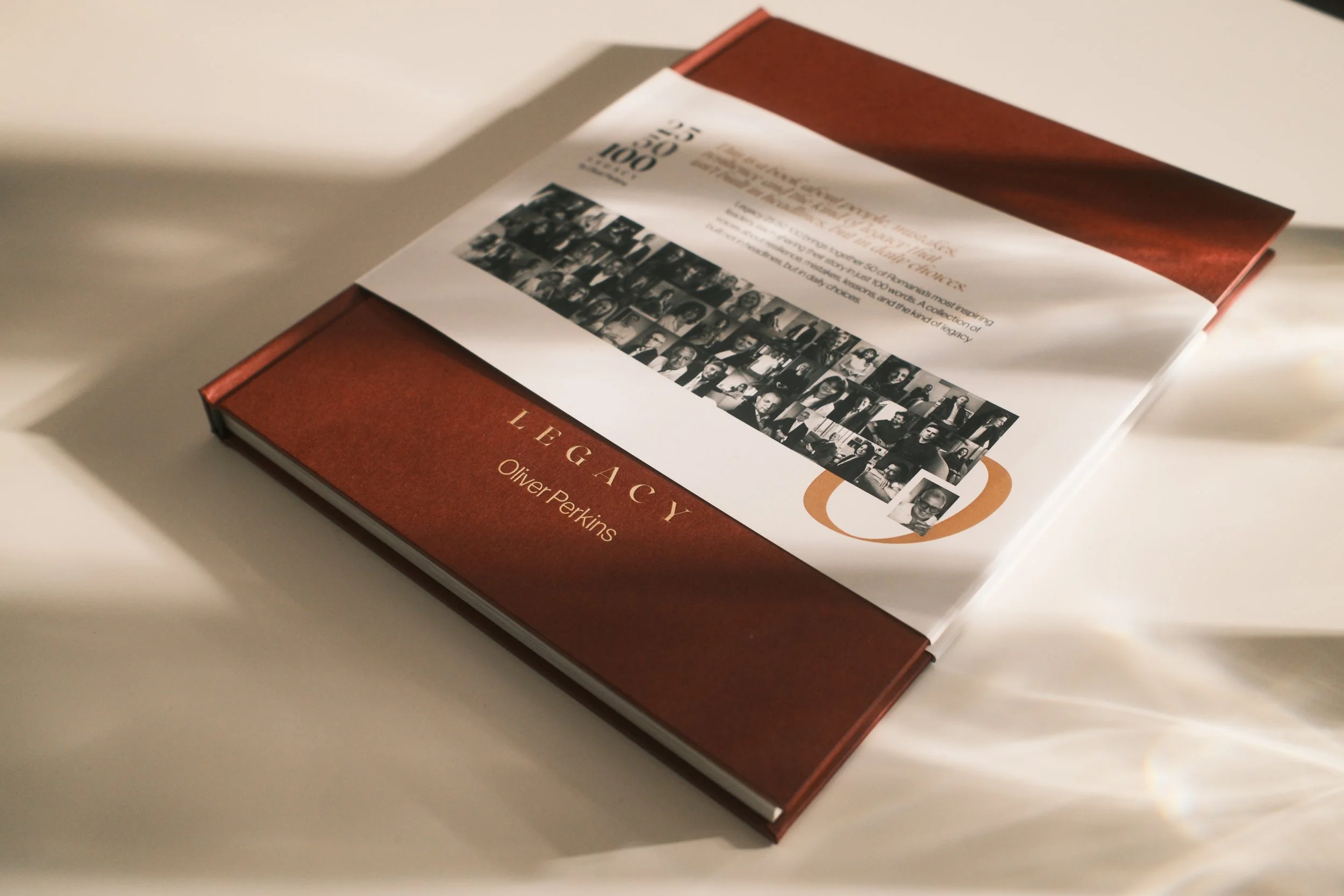 A hardcover book titled 'Legacies' by Oliver Perkins, lying on a white surface with a shadow cast beside it, featuring a collage of black-and-white photographs on the cover.