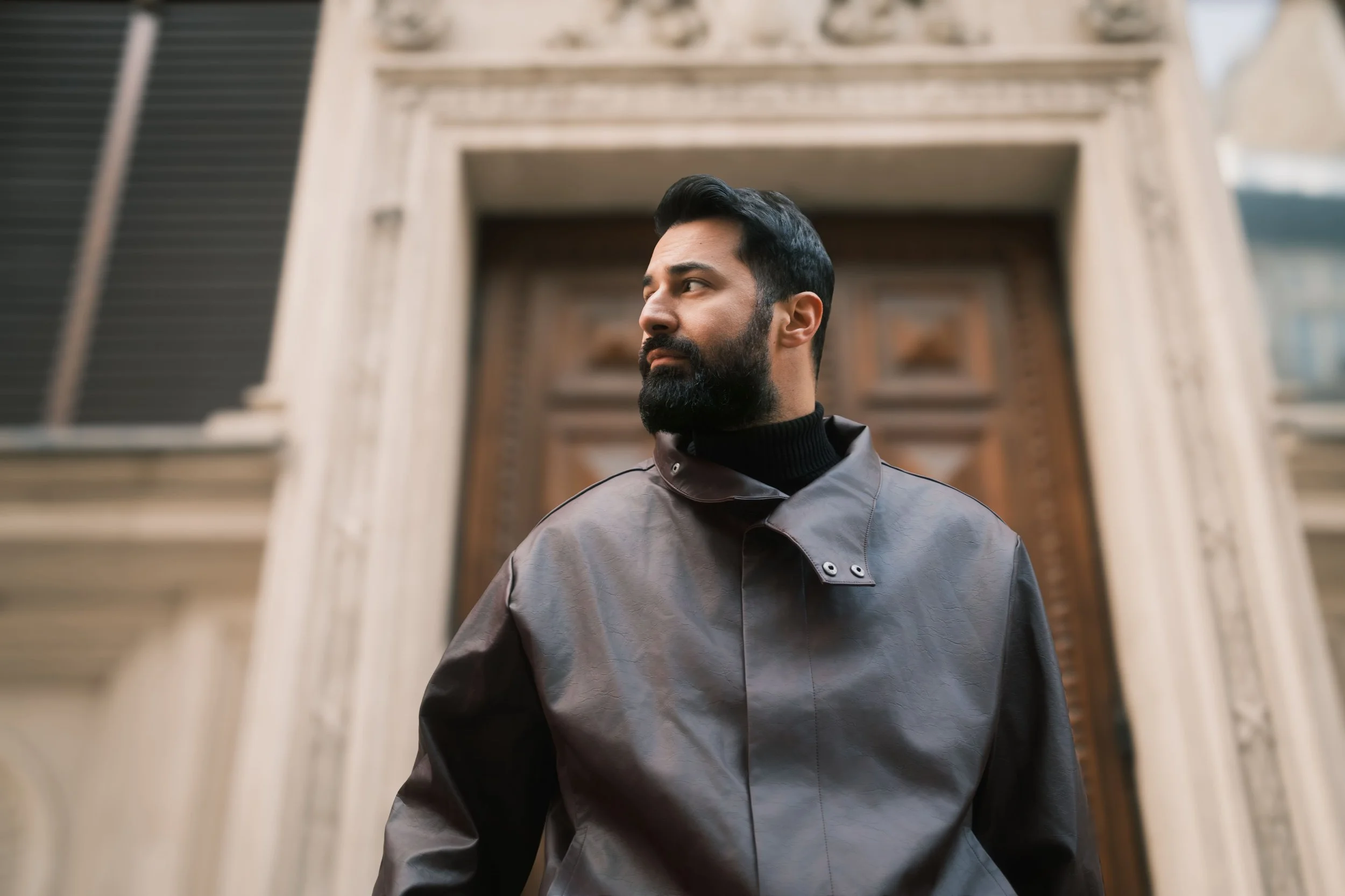 A portrait of Ahmed Hatem in front of an ornate wooden door, wearing a dark gray jacket and black turtleneck, framed by classical details.
