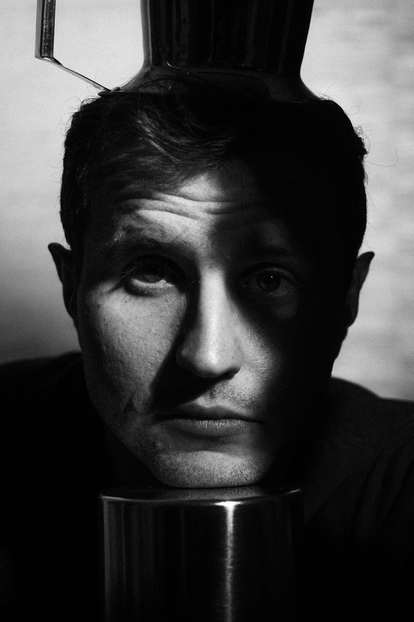 Black and white photo of a man with a mug on his head, resting his face on a reflective surface.