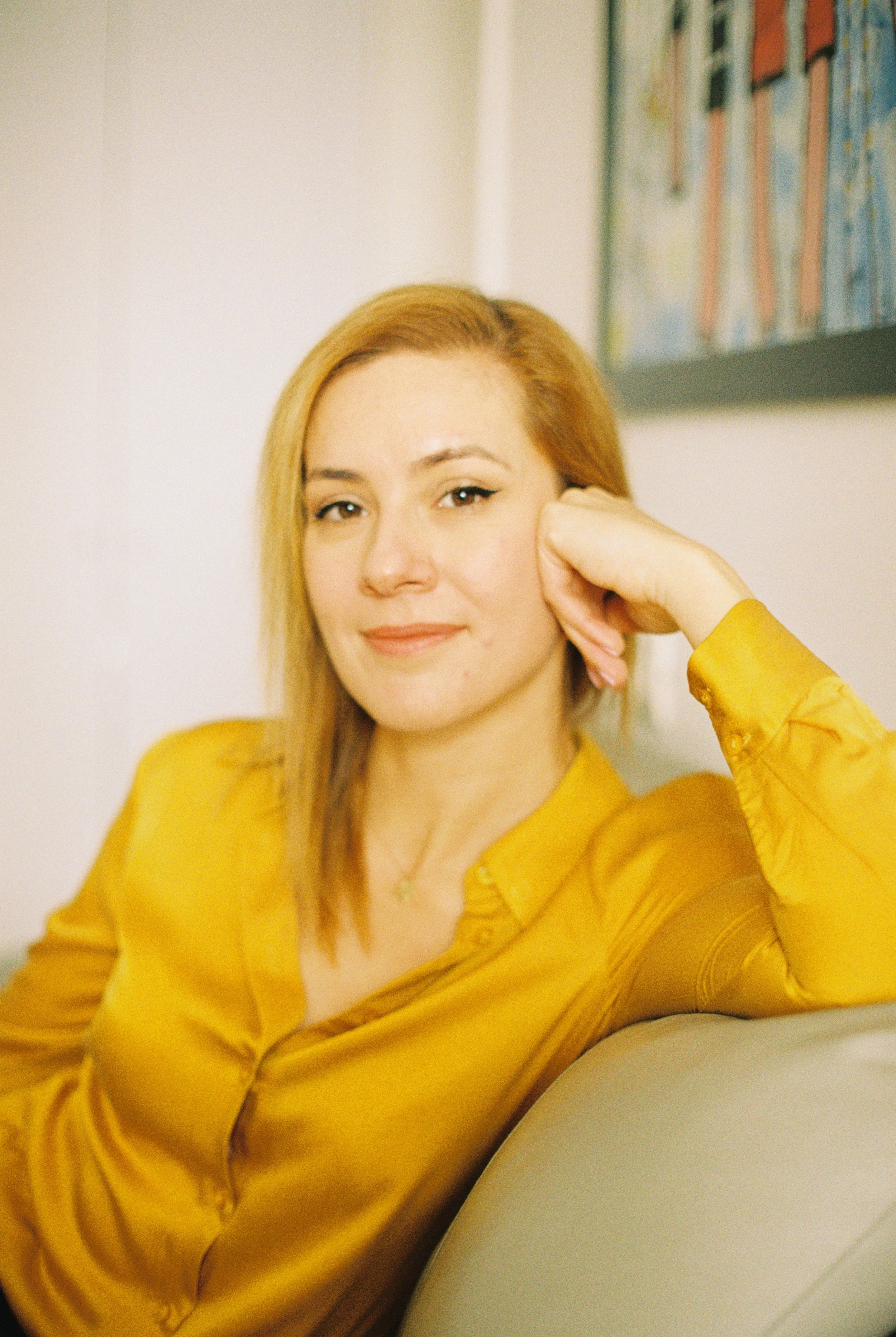 A woman with blond hair and light skin wearing a yellow satin blouse, sitting on a light-colored sofa, smiling slightly, with her head resting on her hand, in front of a bookshelf.