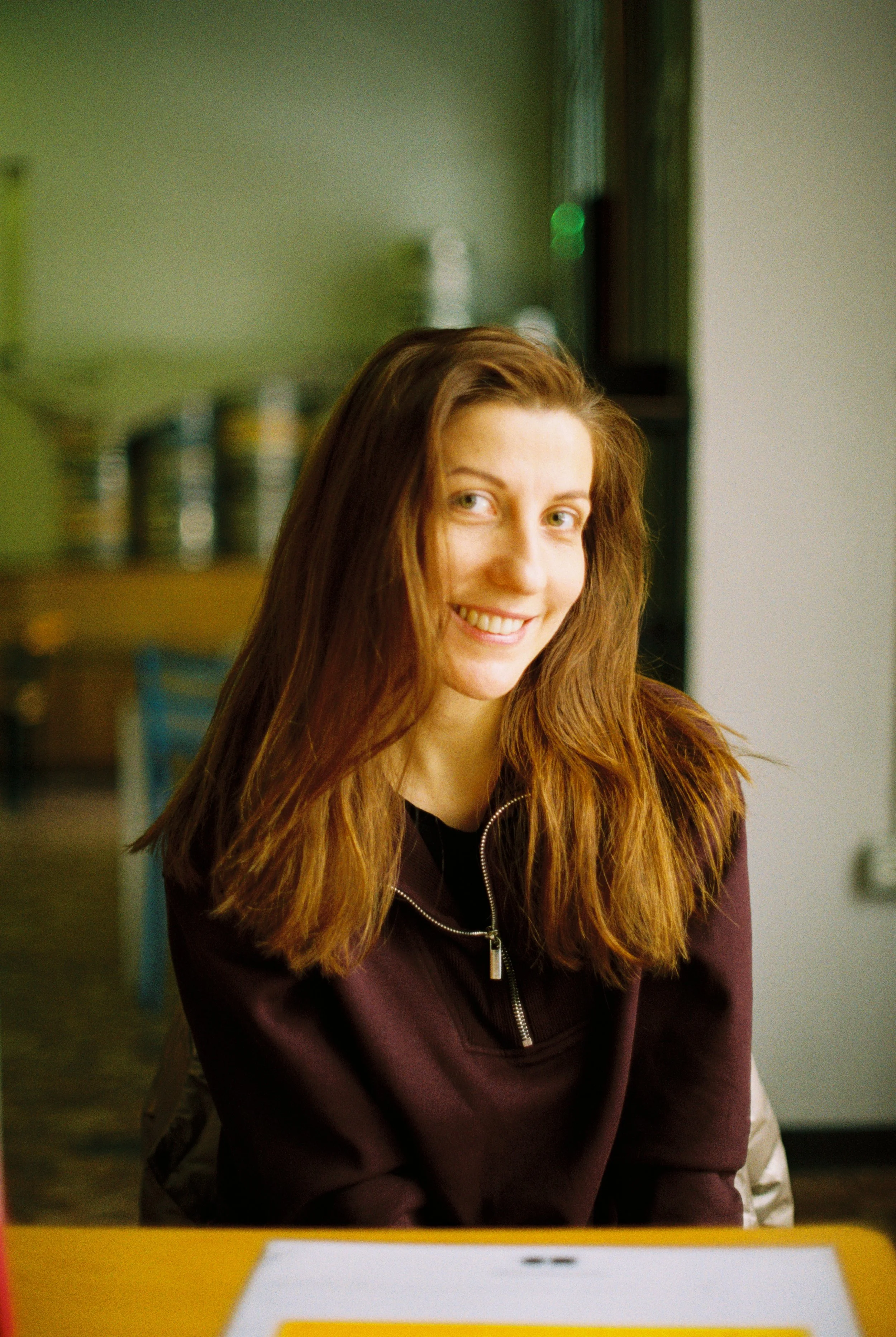 A young woman with long, wavy red hair, smiling and looking at the camera, sitting at a table with a yellow surface in a room with colorful shelves in the background.