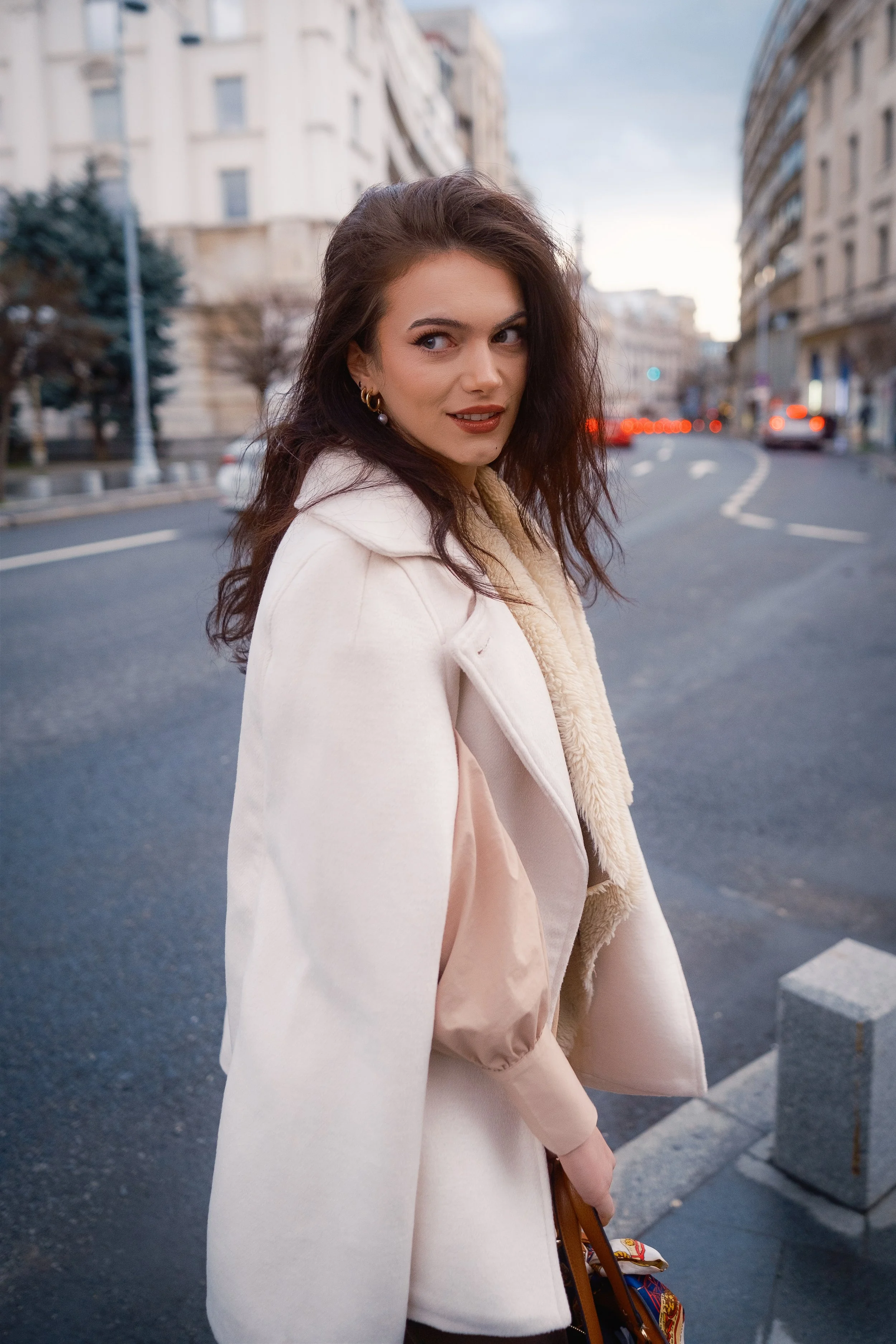 A woman with wavy, dark hair styled in loose waves, wearing a cream-colored coat with a shearling collar, standing on a city street during evening or dusk. She is looking over her shoulder with a slight smile, with buildings and blurred car lights in