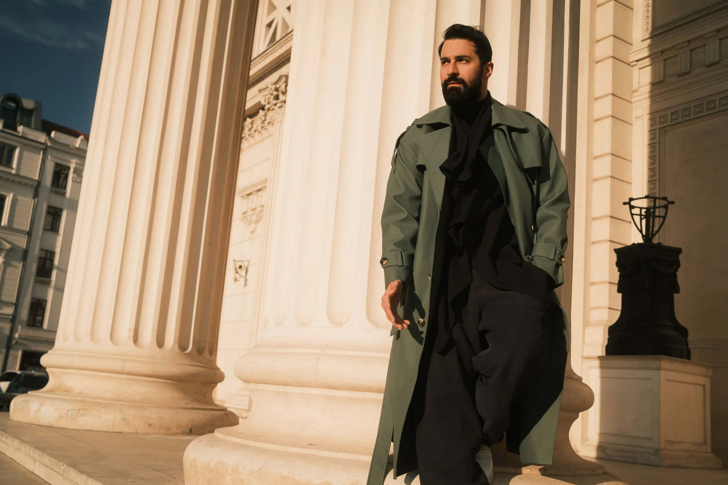 Ahmed Hatem leans against a stone column in Bucharest, wearing a green trench coat over black clothing. Soft natural light creates a calm, cinematic portrait.