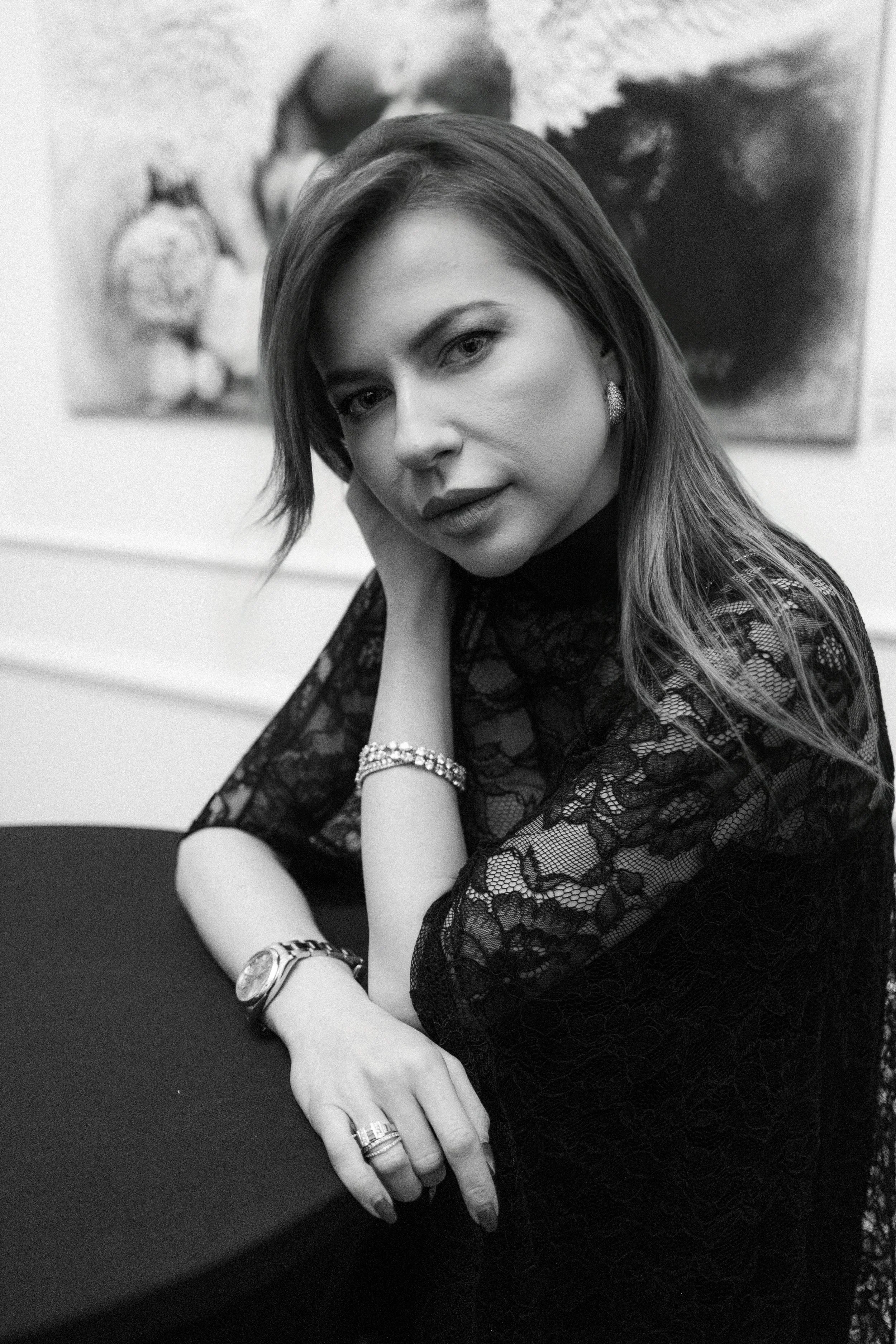 Black and white portrait of a woman wearing a lace top, with jewelry, sitting at a table, with artwork in the background.
