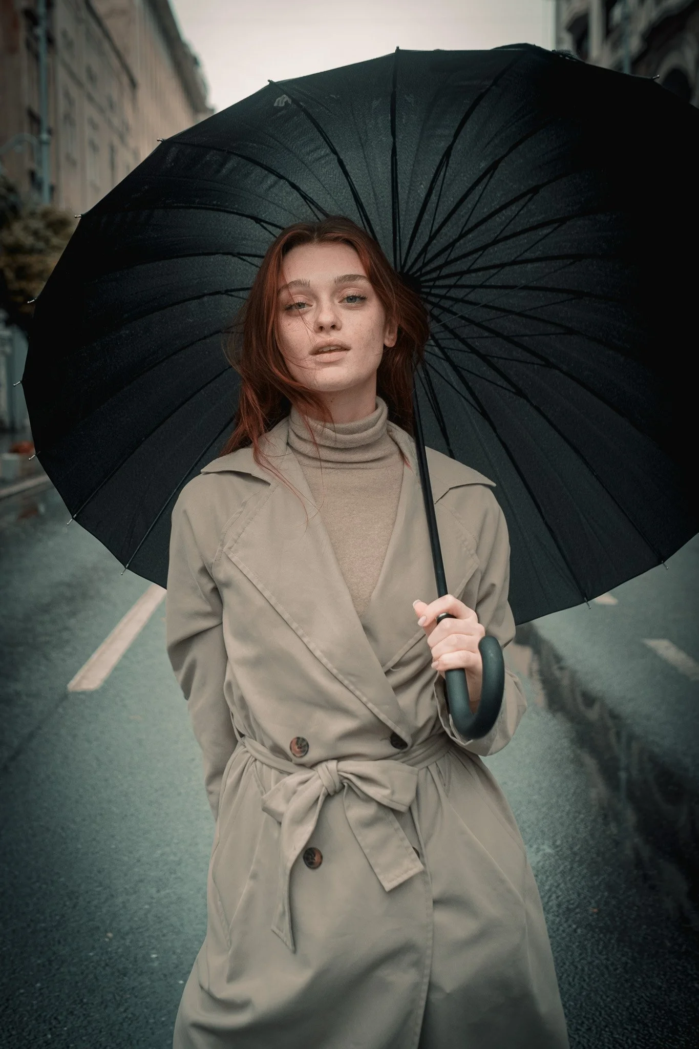 A young woman with red hair holding a black umbrella stands on a rain-soaked city street during overcast weather, wearing a beige trench coat and turtleneck.