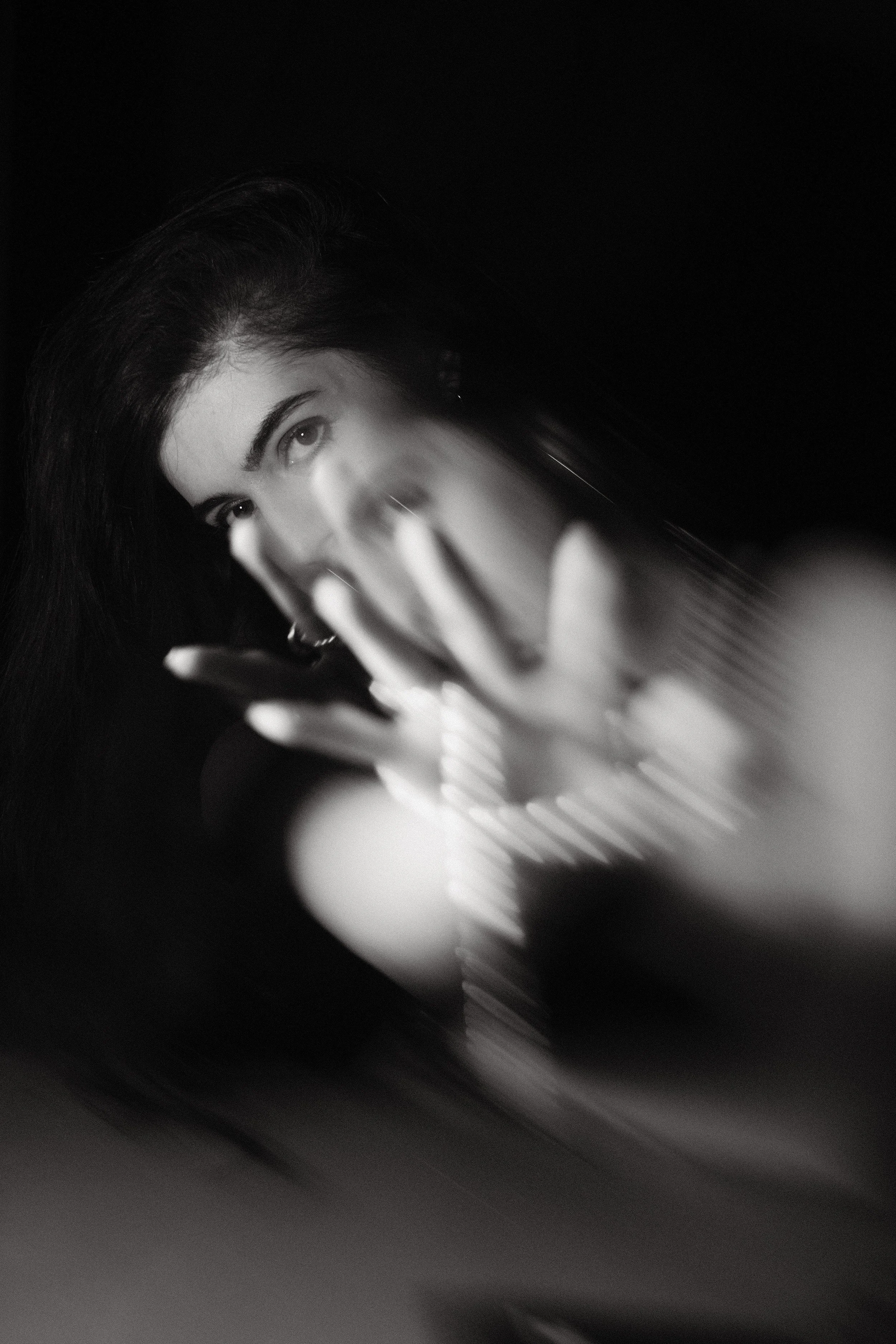 Black and white close-up of a woman with dark hair, with her hand partially covering her face, against a dark background.