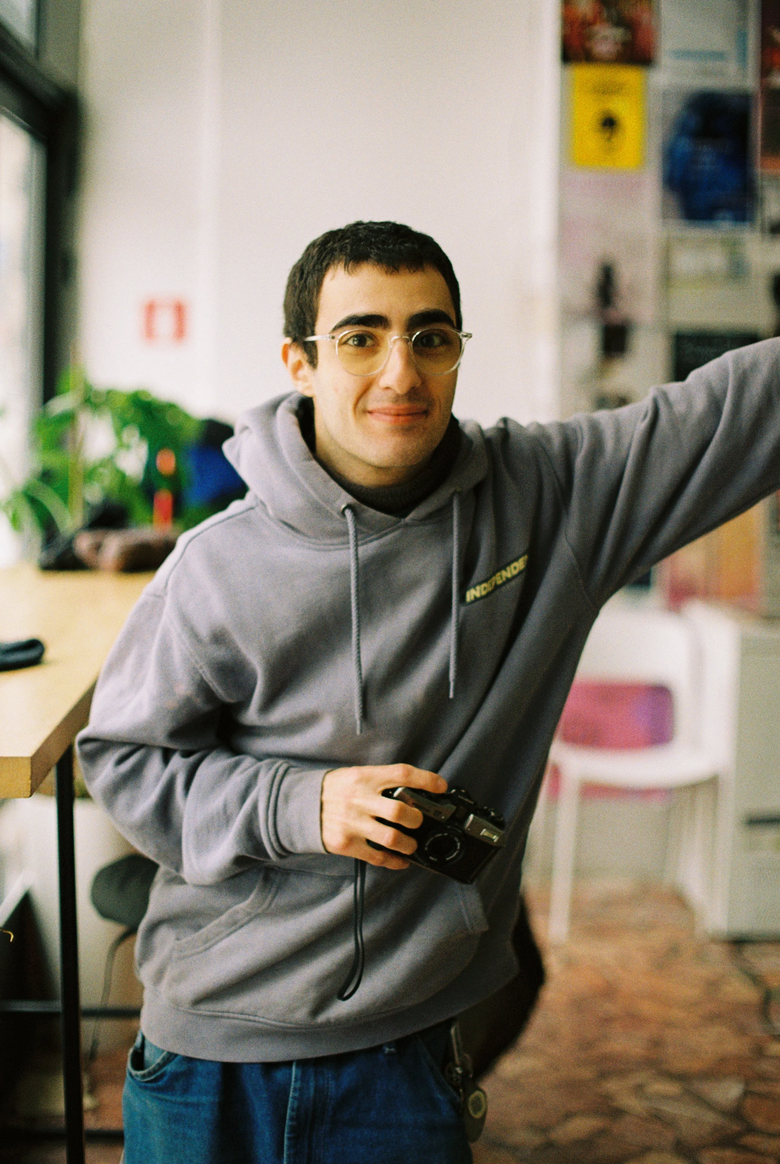A young man with glasses holding a camera, wearing a gray hoodie, in an indoor setting with colorful posters and a white chair in the background.