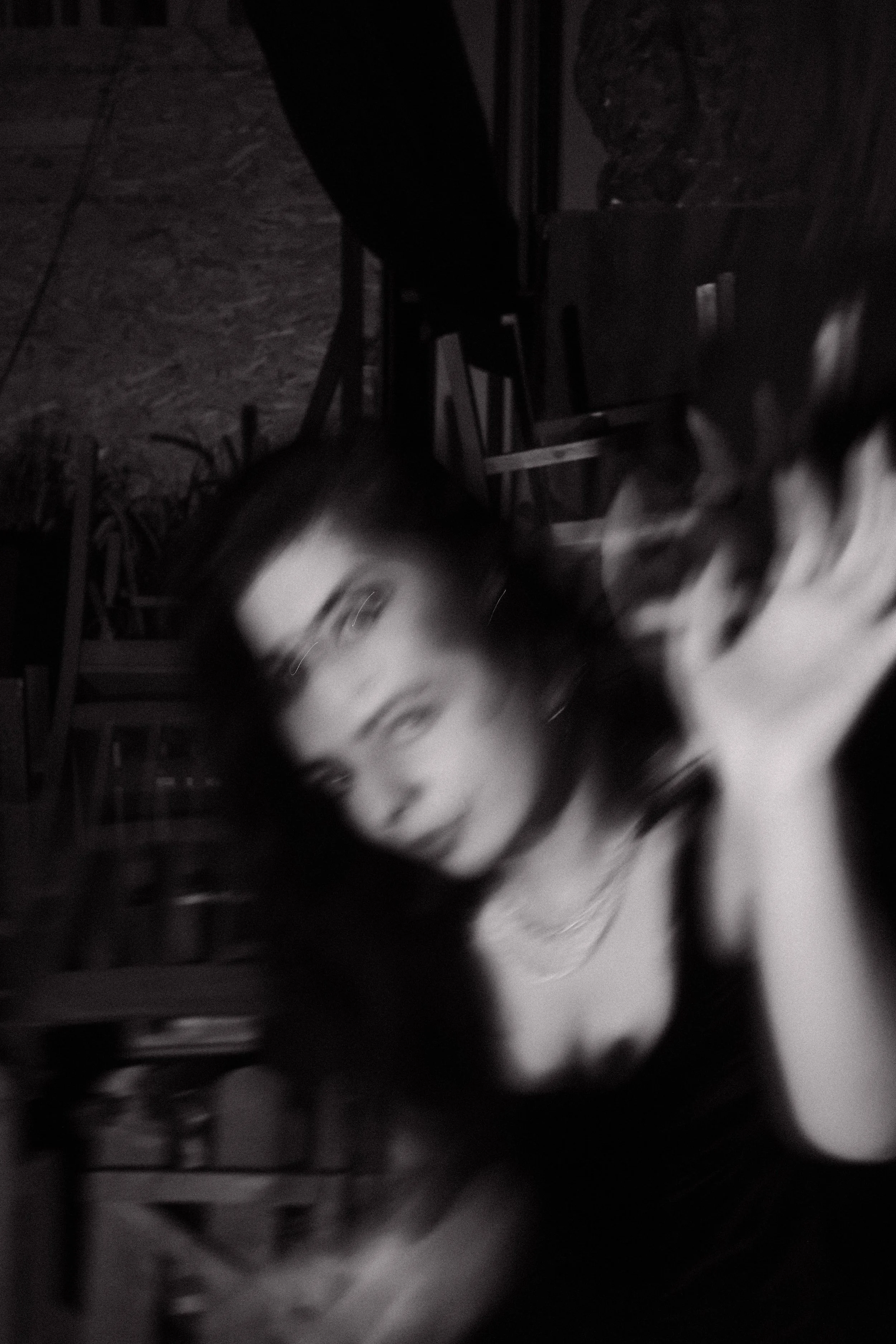 A blurry black-and-white photo of a person's face and upper body, with long hair, in a dimly lit room with chairs and furniture in the background.