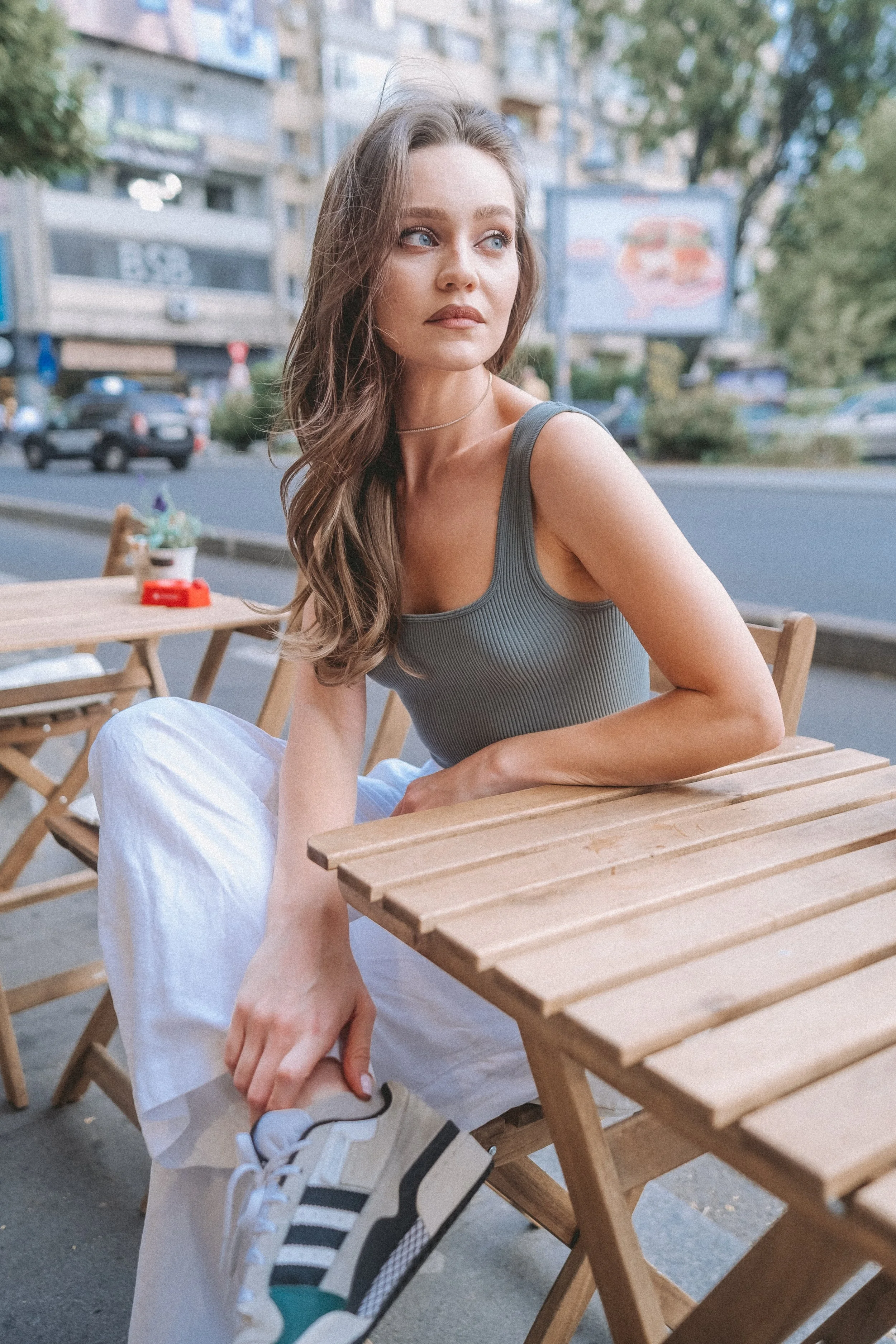 A young woman with long wavy brown hair and blue eyes sitting outdoors at a wooden table on a city street. She is wearing a gray tank top and white pants, looking away contemplatively.