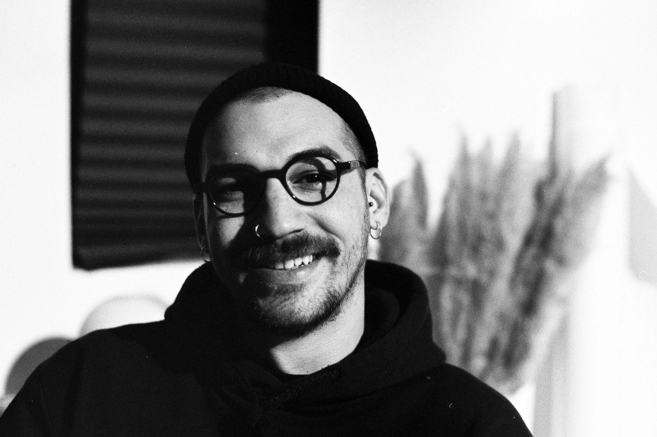 Black and white photo of a smiling man with beard and glasses, wearing a hoodie and beanie, indoors with some abstract decorations in the background.
