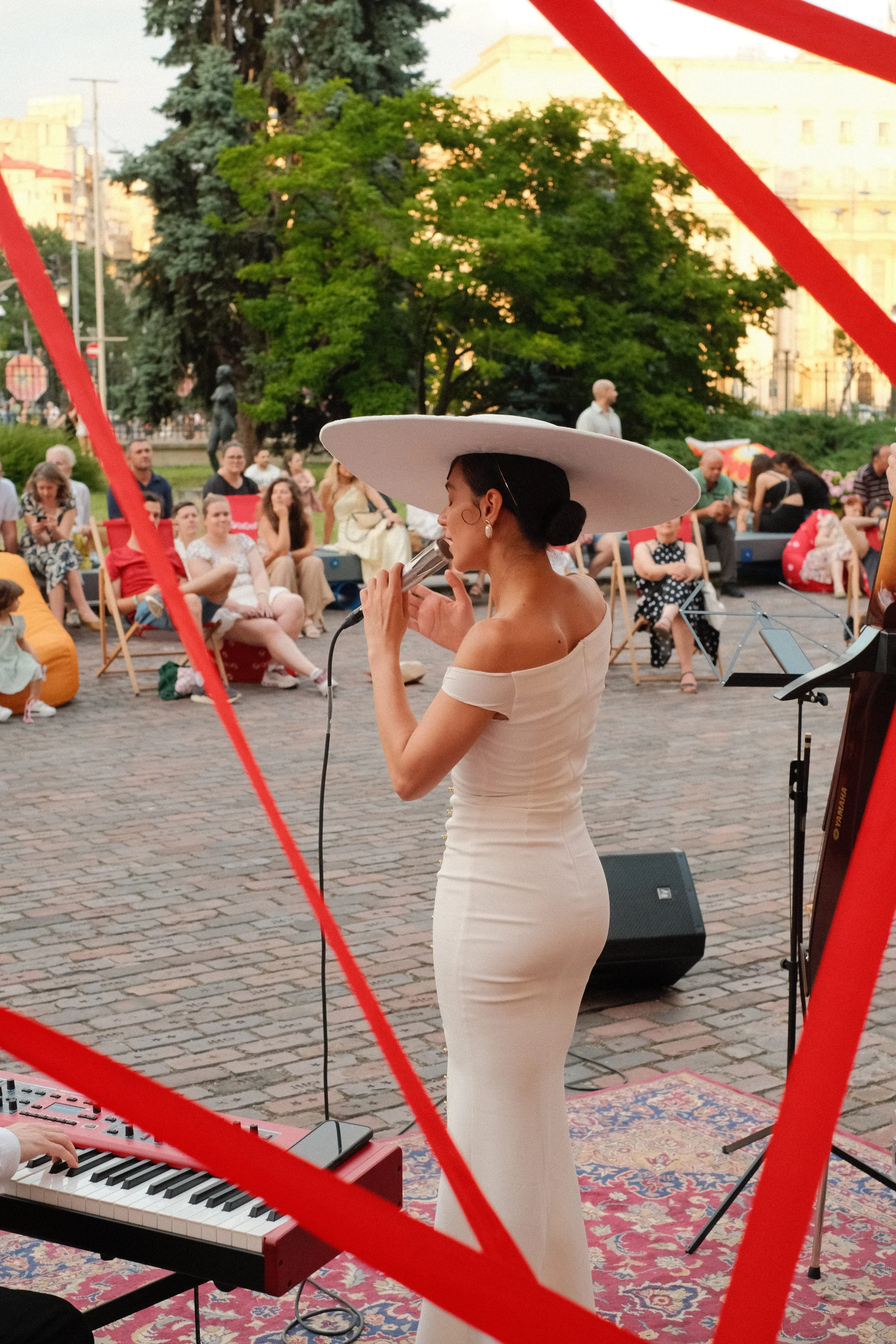 A woman in a white dress and large white hat singing into a microphone during a live outdoor music performance, with an audience seated on chairs and benches in a park-like setting.