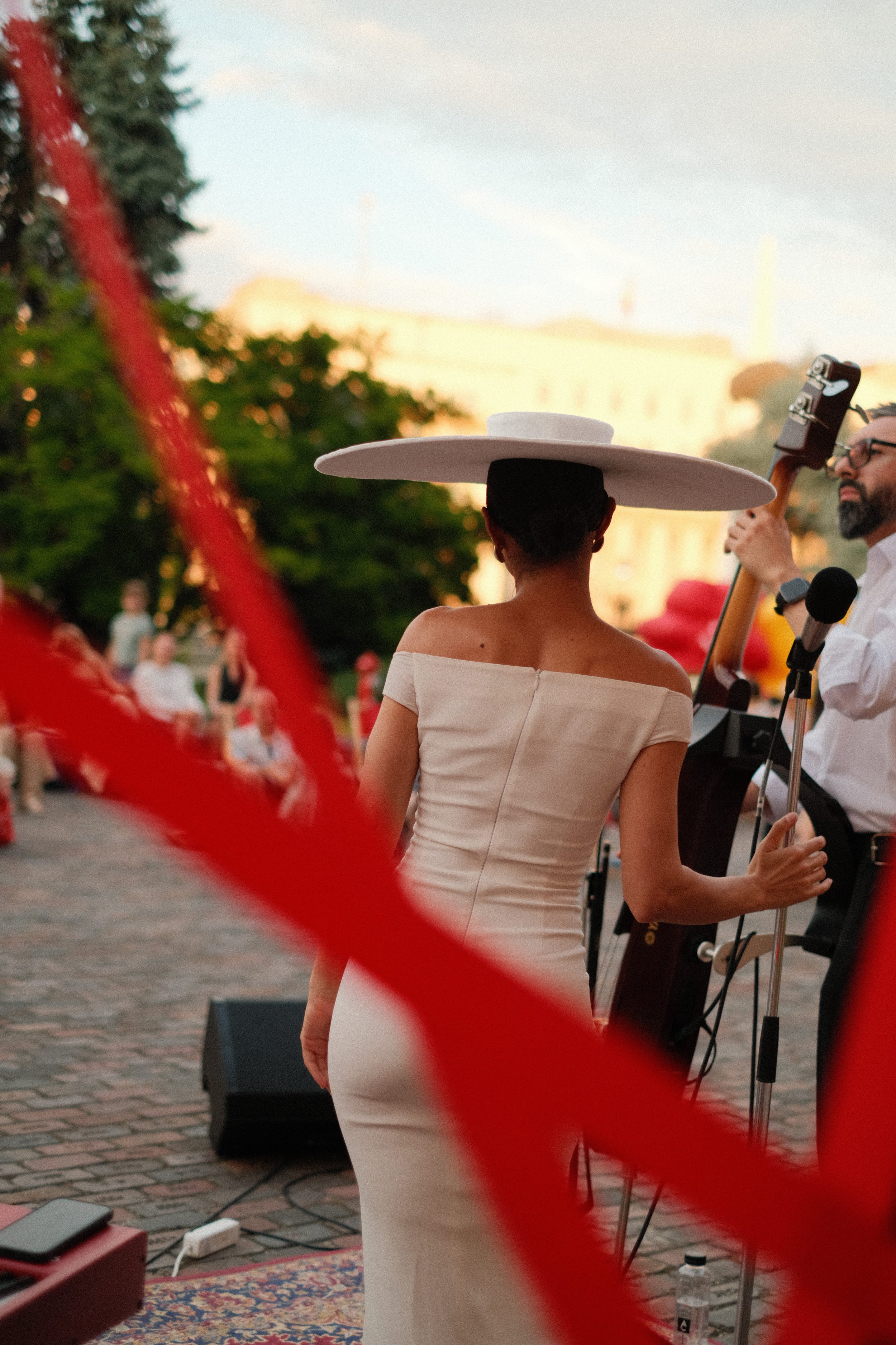 A woman in a white dress and a large white hat stands outdoors during a live music performance, with a man playing guitar and another man with glasses also performing. Audience members are visible in the background.