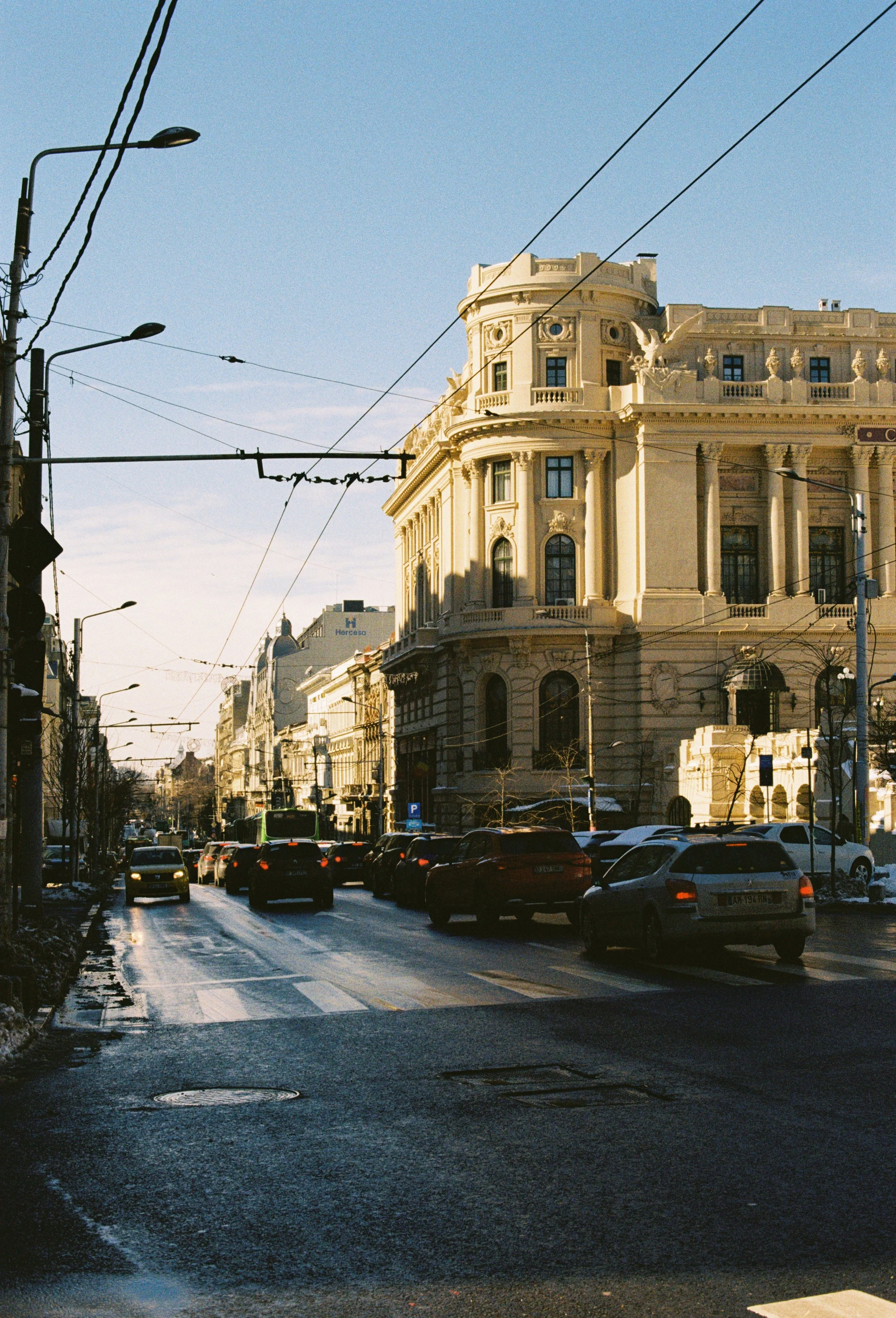 City street scene with historic cream-colored building featuring columns and decorative architecture, cars parked along the street, overhead tram wires, and a clear blue sky.