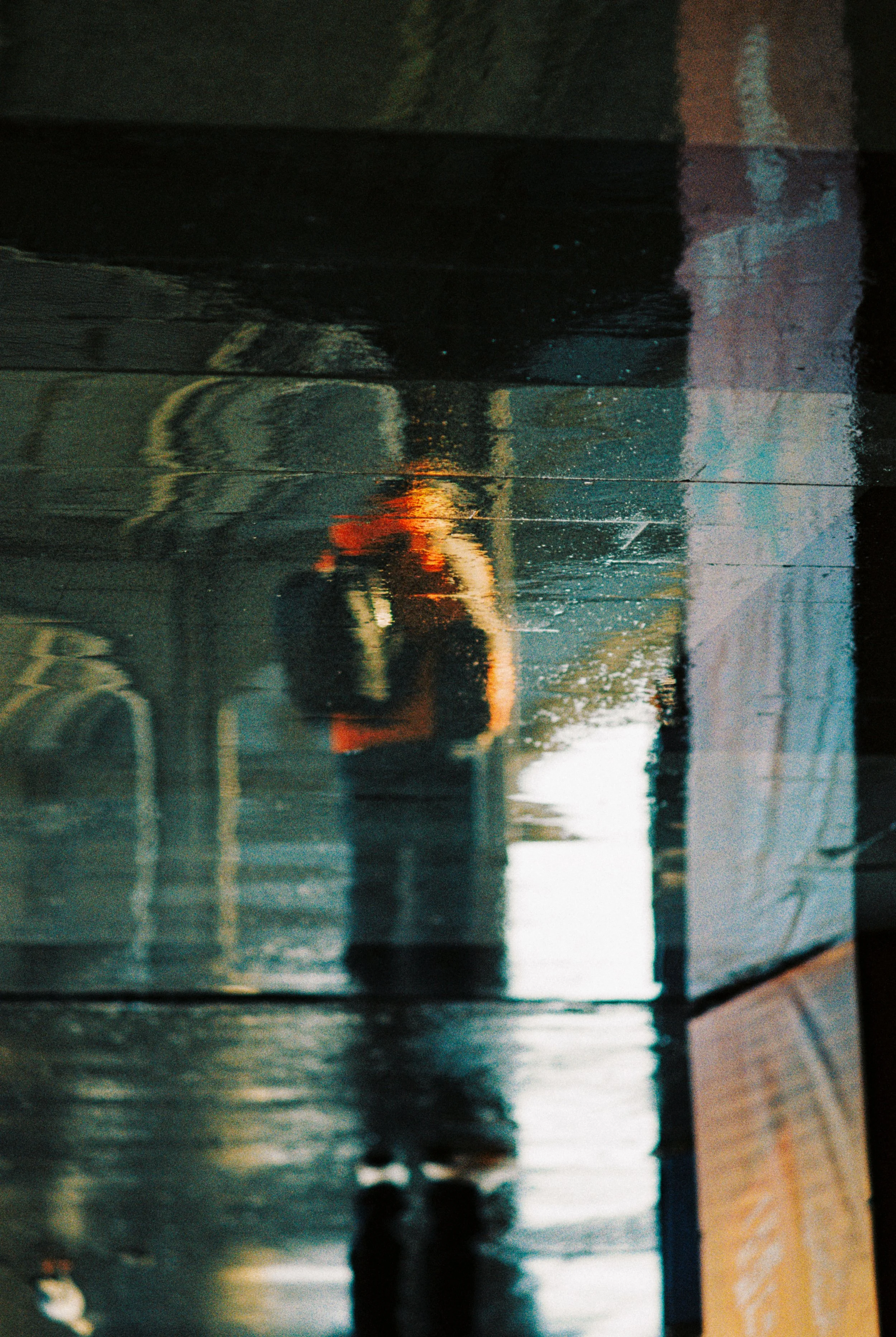 A person taking a selfie in a wet, reflective surface, possibly a puddle or shiny floor, with buildings and sky reflected around them.