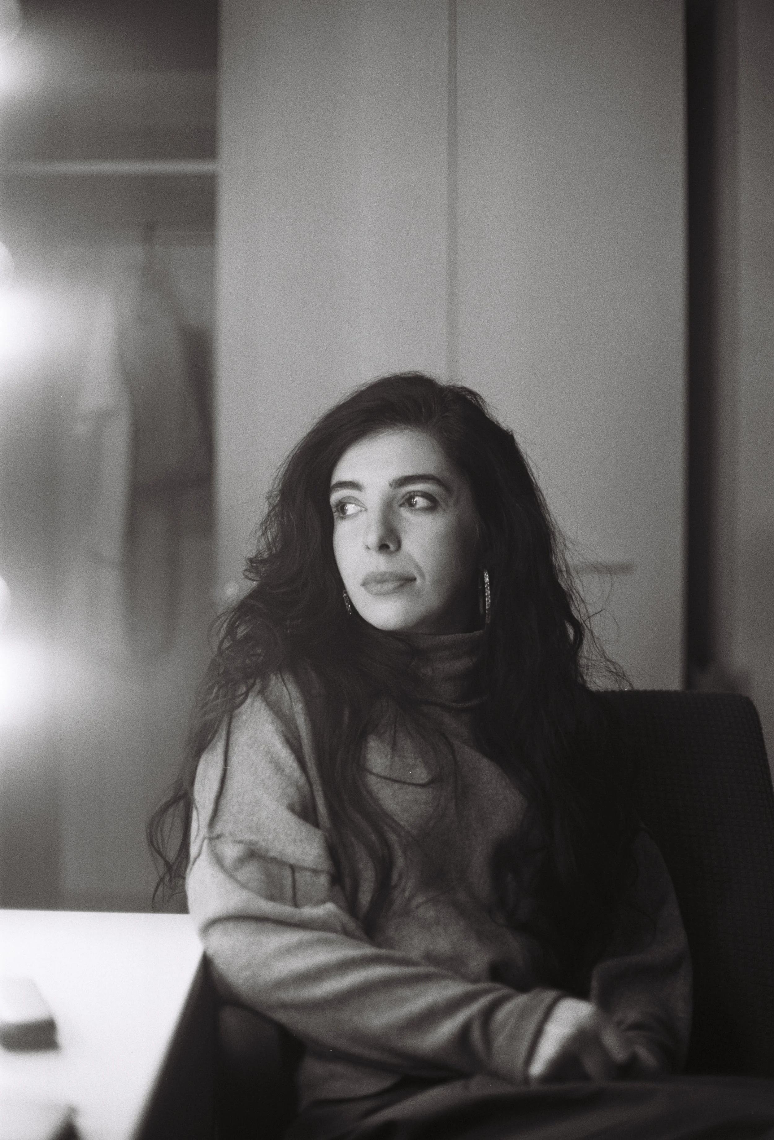 A woman with long, wavy dark hair, sitting indoors, looking to her right with a thoughtful expression, wearing earrings and a hoodie.
