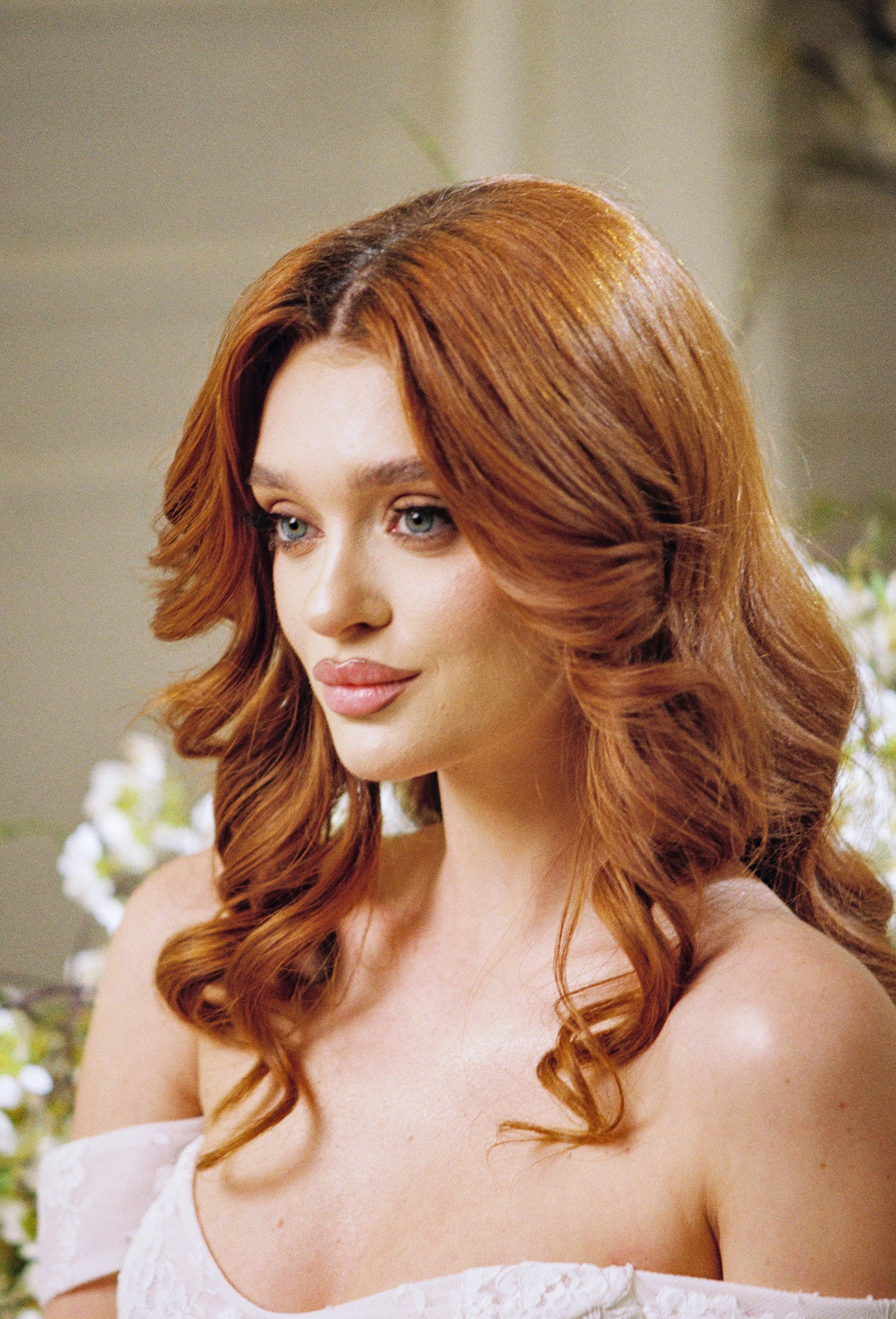 A woman with wavy red hair and blue eyes, wearing a white off-shoulder top, looking thoughtfully to the side, with a blurred background of white flowers and greenery.