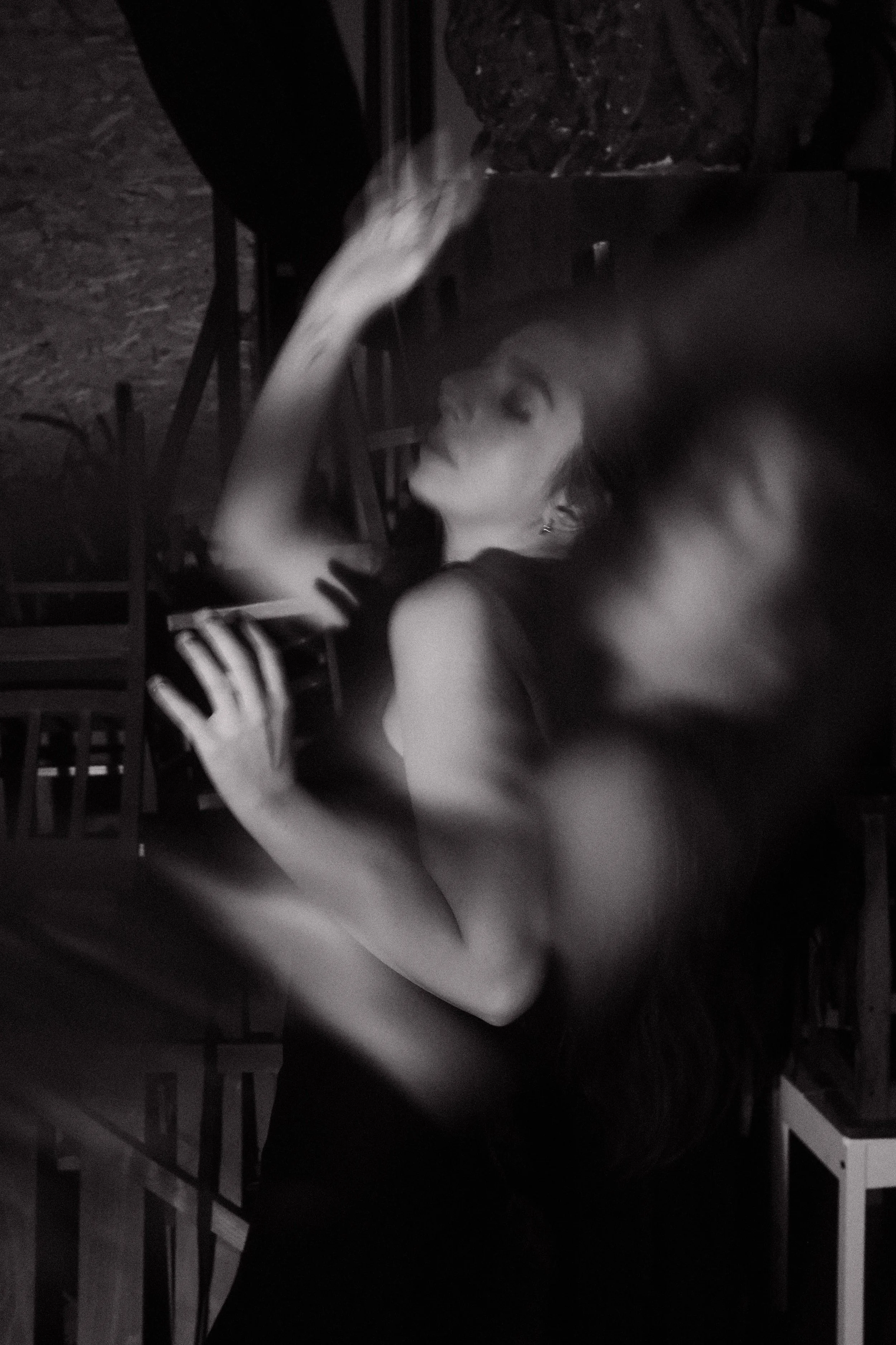A woman with short hair and earrings is sitting on a chair, holding and looking at a smartphone. She is in a dimly lit environment, and part of her arm is blurred due to movement.