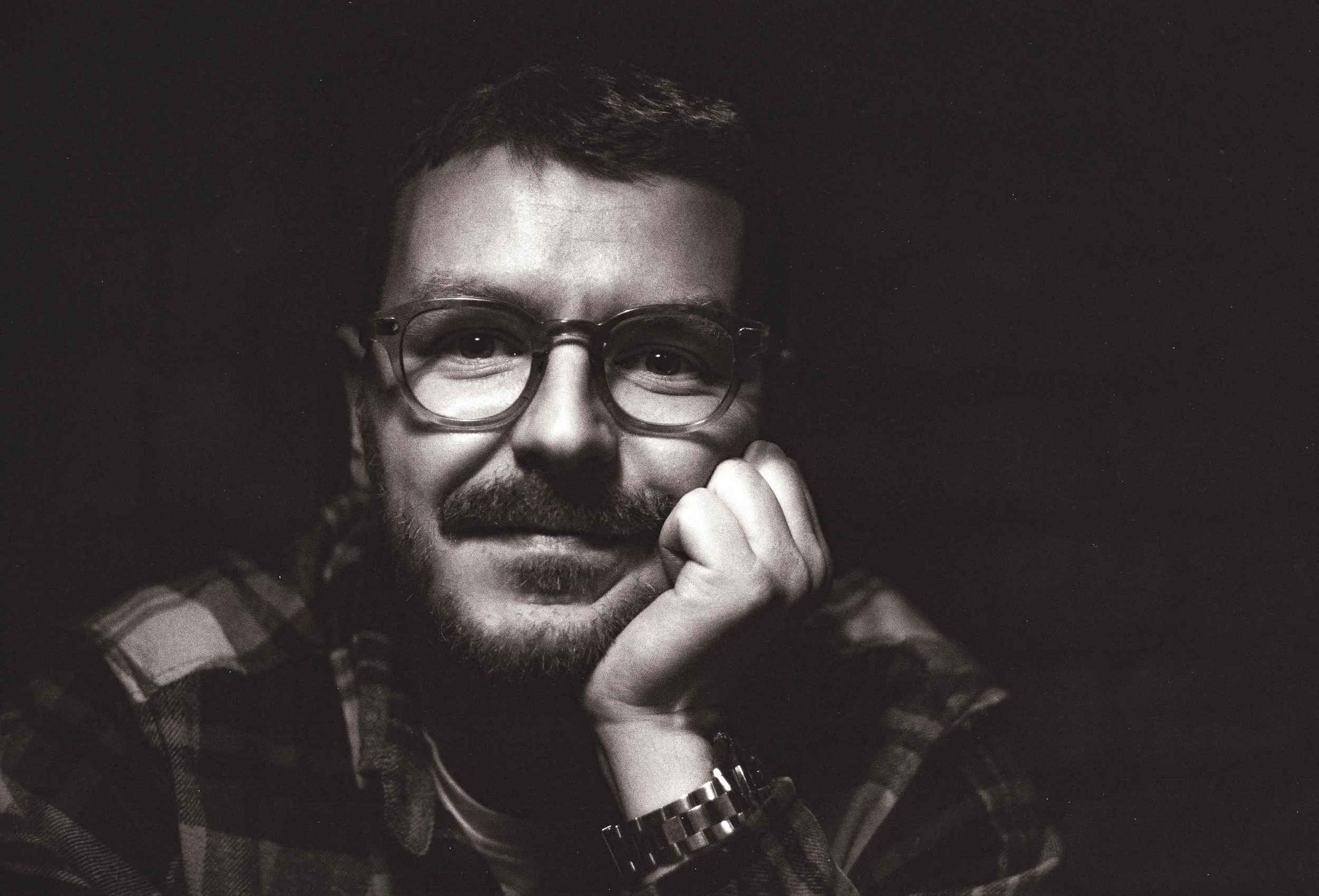 A black-and-white portrait of a man with glasses and a beard, resting his chin on his hand, looking at the camera.
