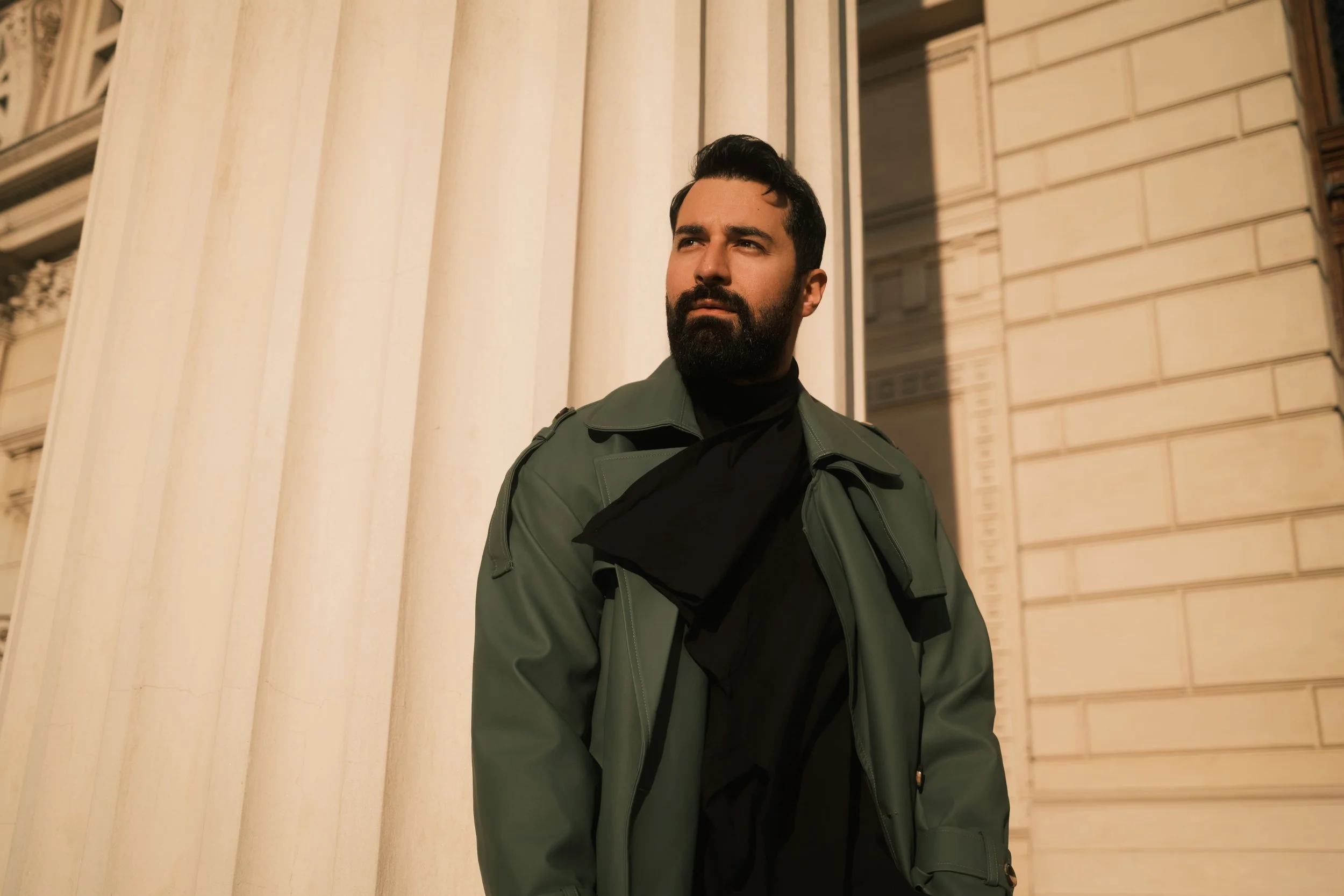 A portrait of Ahmed Hatem framed by tall architectural columns, dressed in black and a green jacket, shaped by natural daylight and structure.