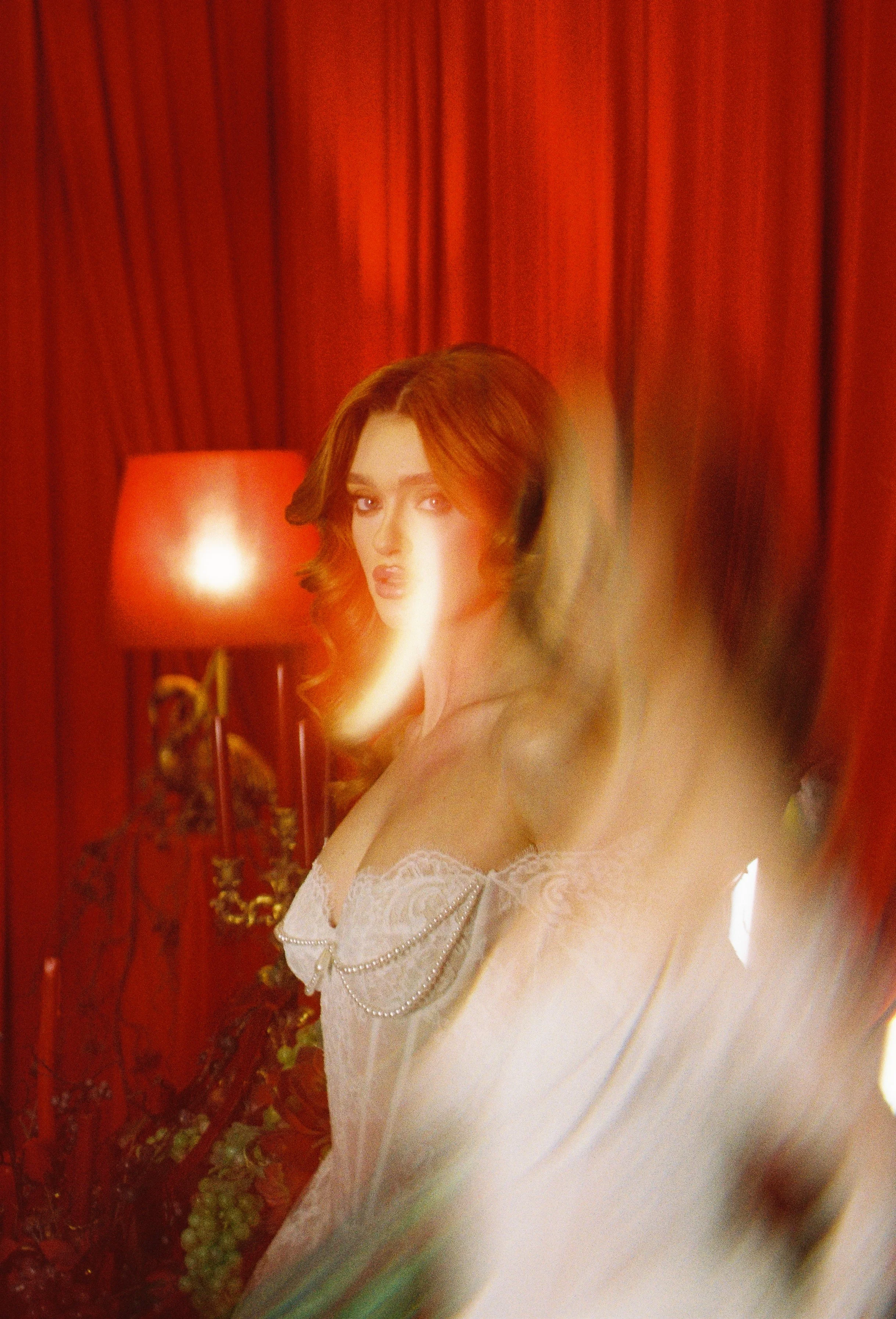 A woman with long reddish-brown hair makes a fashion pose in a room with red curtains, a red lamp, and a blurred foreground. She wears a white lace corset with pearl accents.