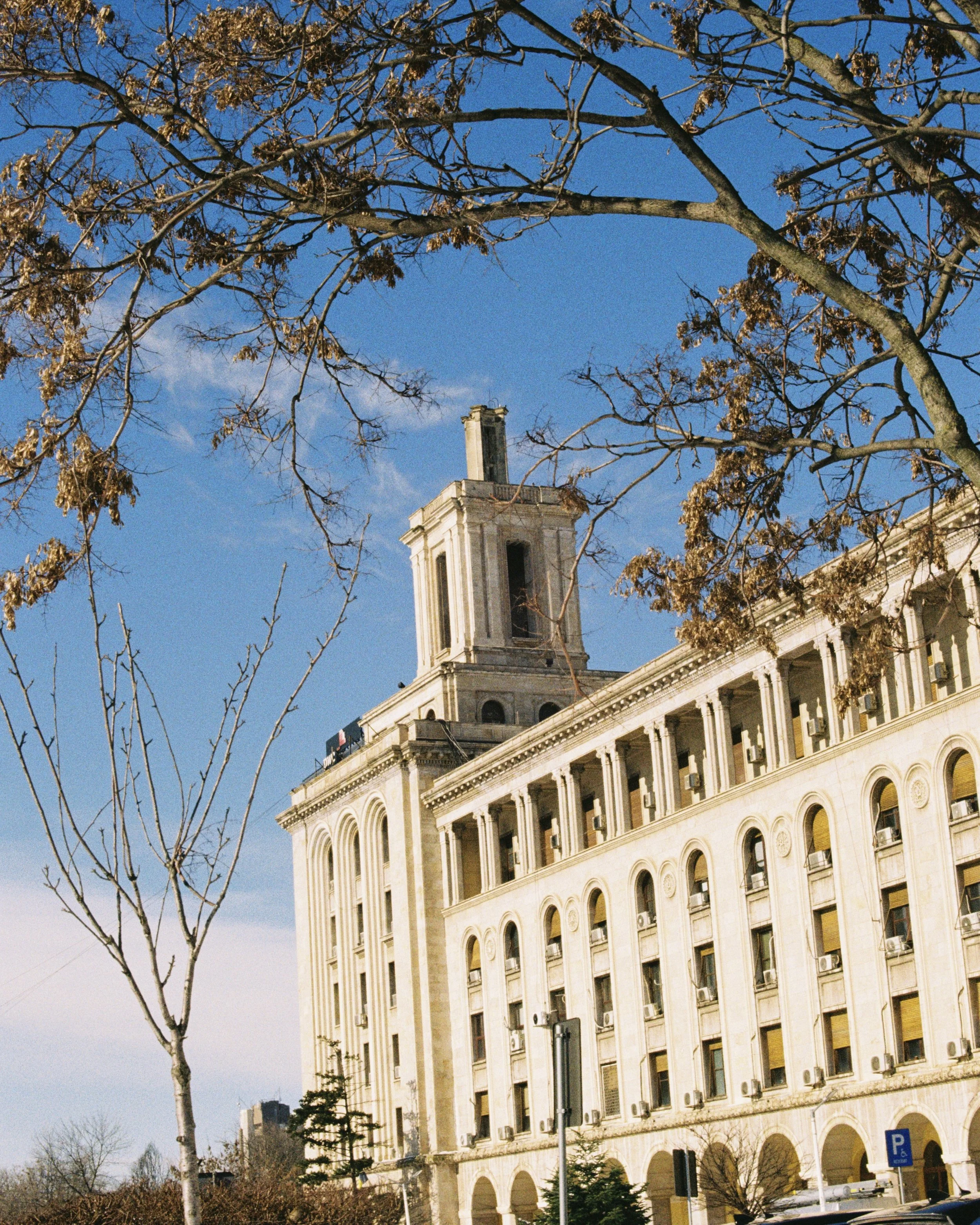 A historic building with classical architecture, featuring arched windows and a tower, under a blue sky with leafless trees in the foreground.