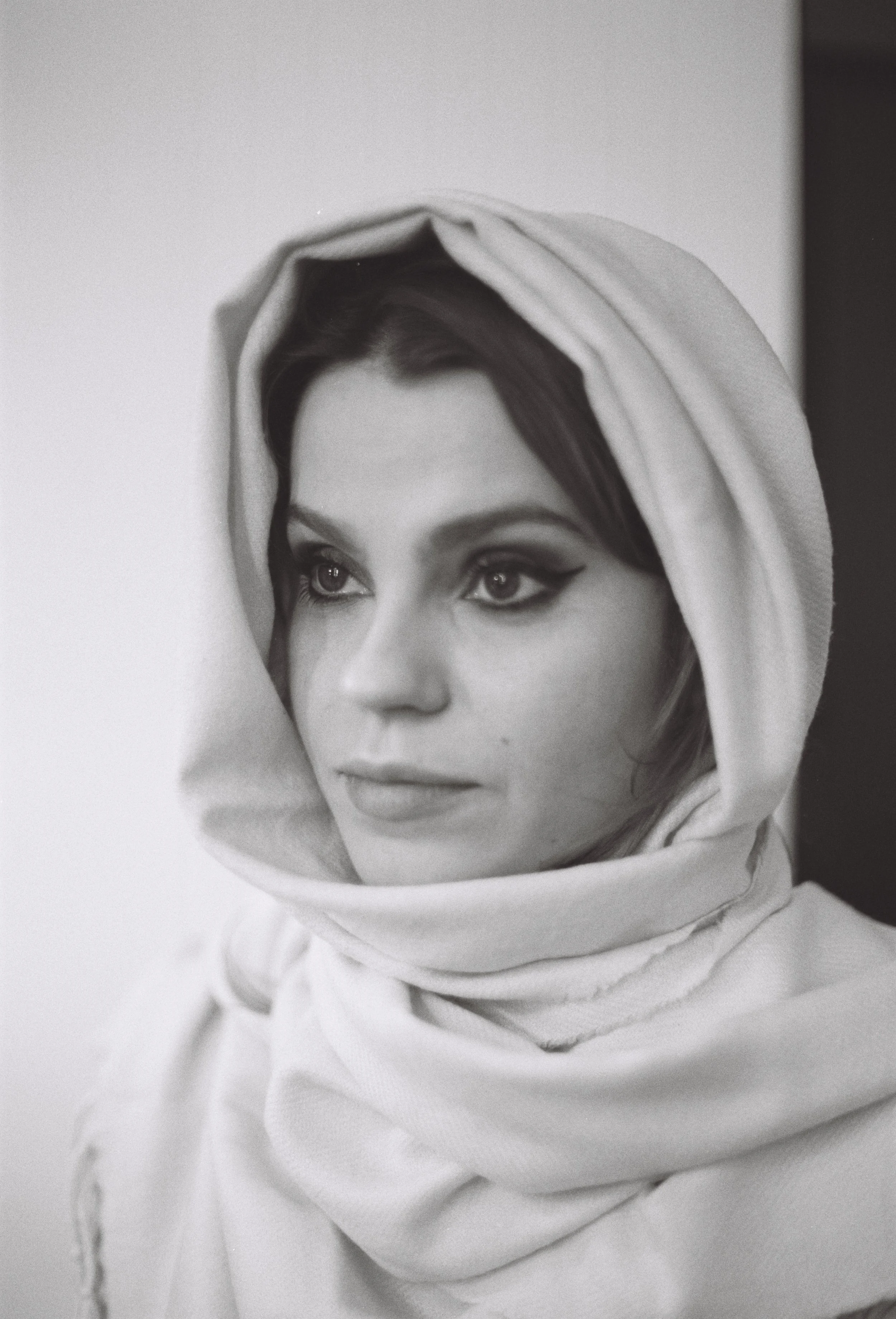A black and white portrait of a woman wearing a light-colored hooded garment, looking to her left with striking eyes and makeup.