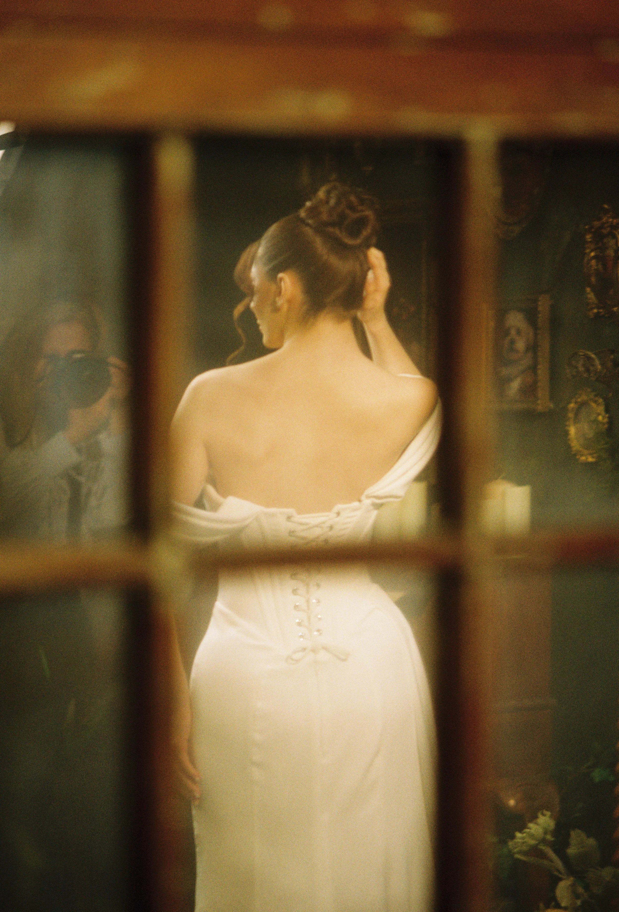 A woman with dark hair styled in an updo, wearing a strapless dress with a corset back, stands in front of a mirror, adjusting her hair.