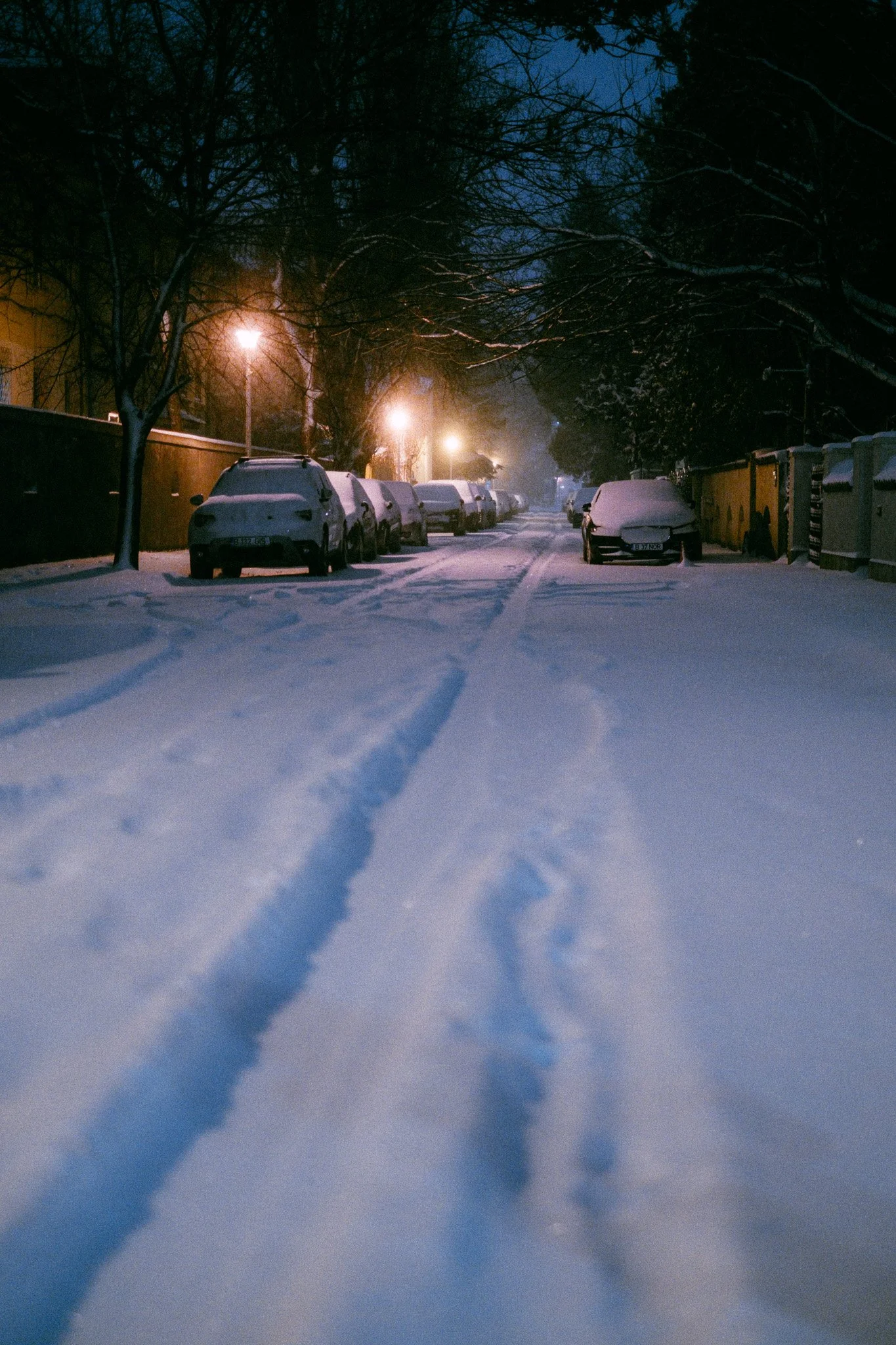 Snow-covered residential street at night with parked cars on both sides and streetlights illuminating the scene.