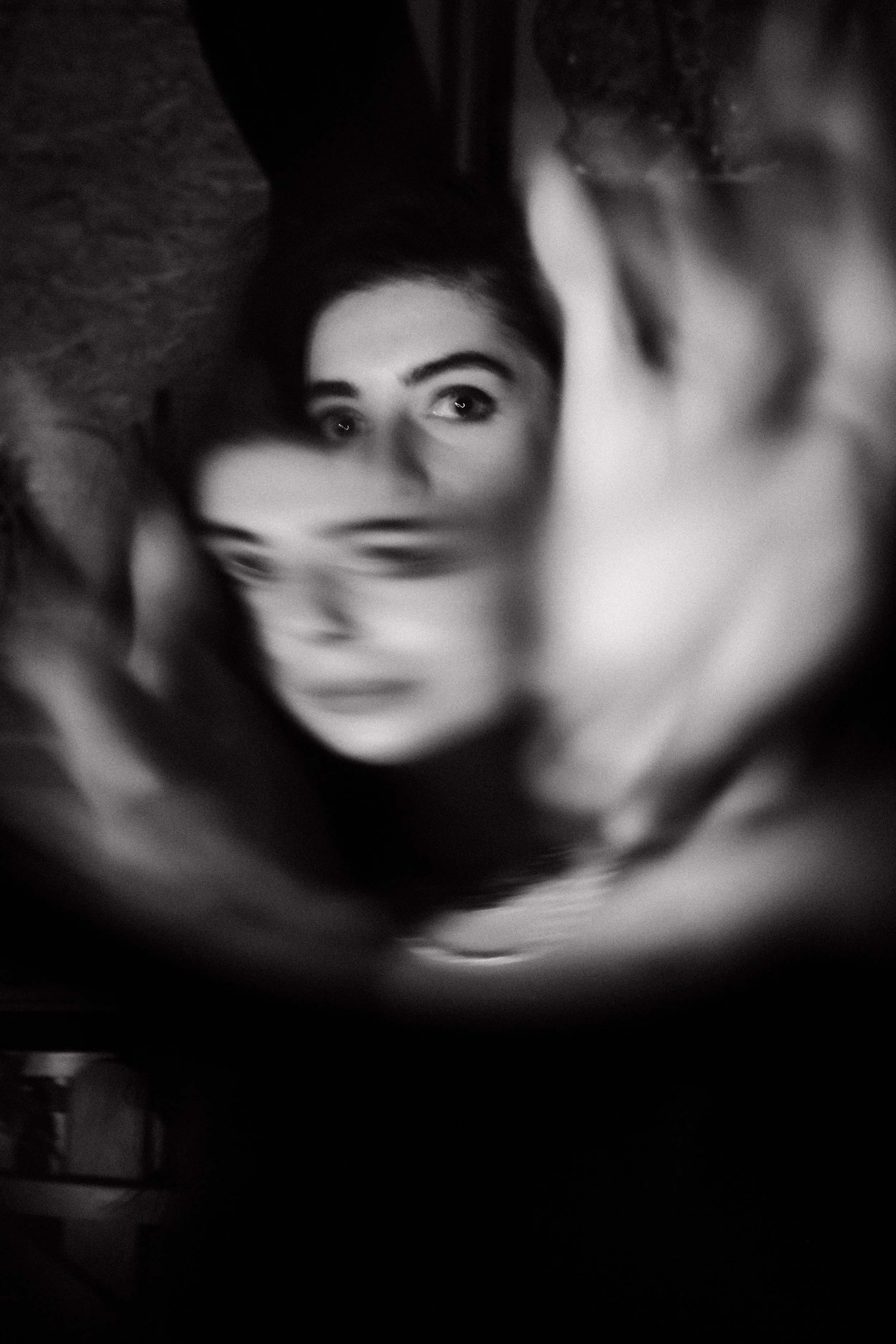 A black and white photo of a woman with dark hair and expressive eyes, facing the camera, with her hands framing her face.