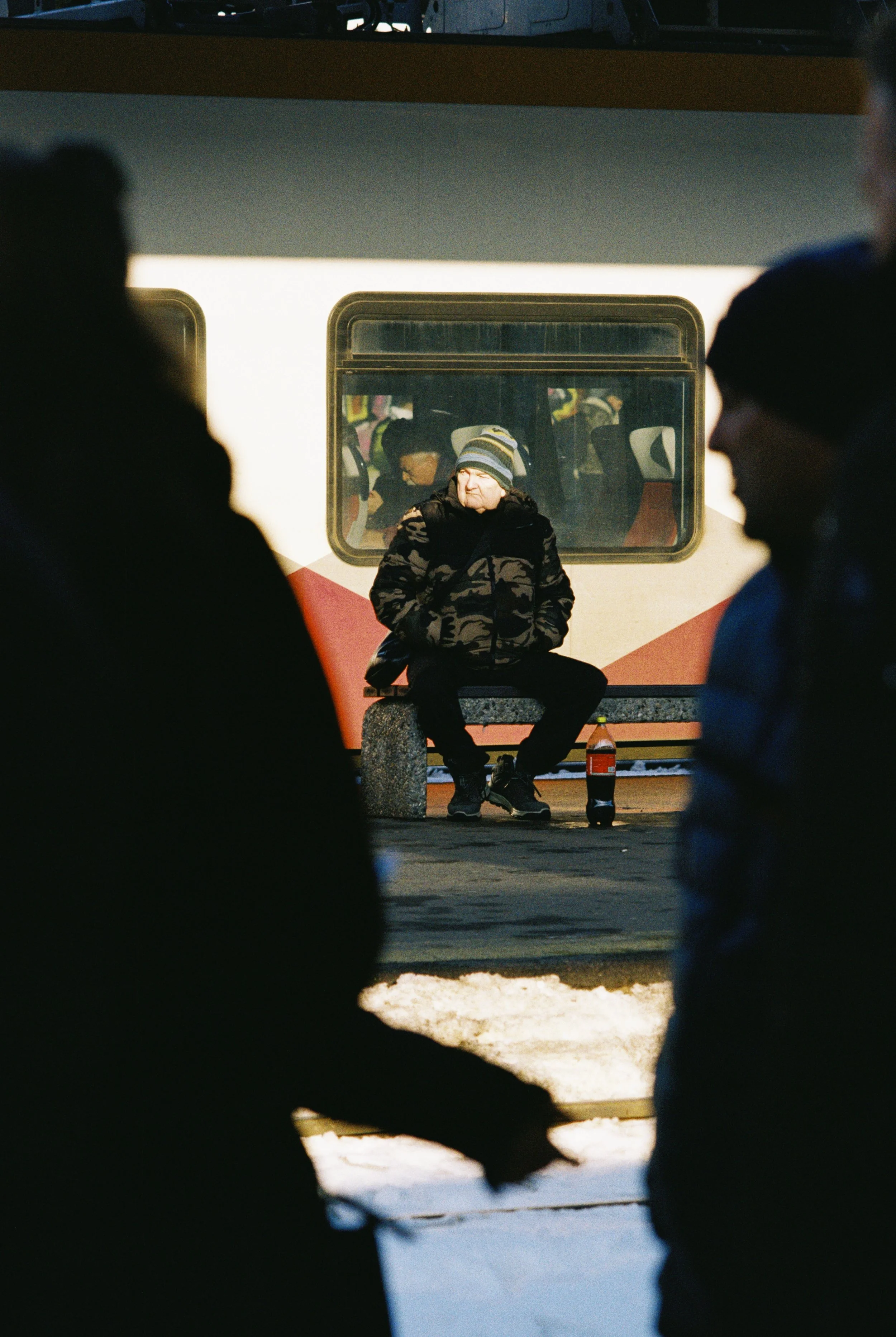 A person wearing a striped beanie and camouflage jacket sitting on a bench at a train station, with a Coca-Cola bottle beside them, framed by silhouetted figures in the foreground and a train in the background.